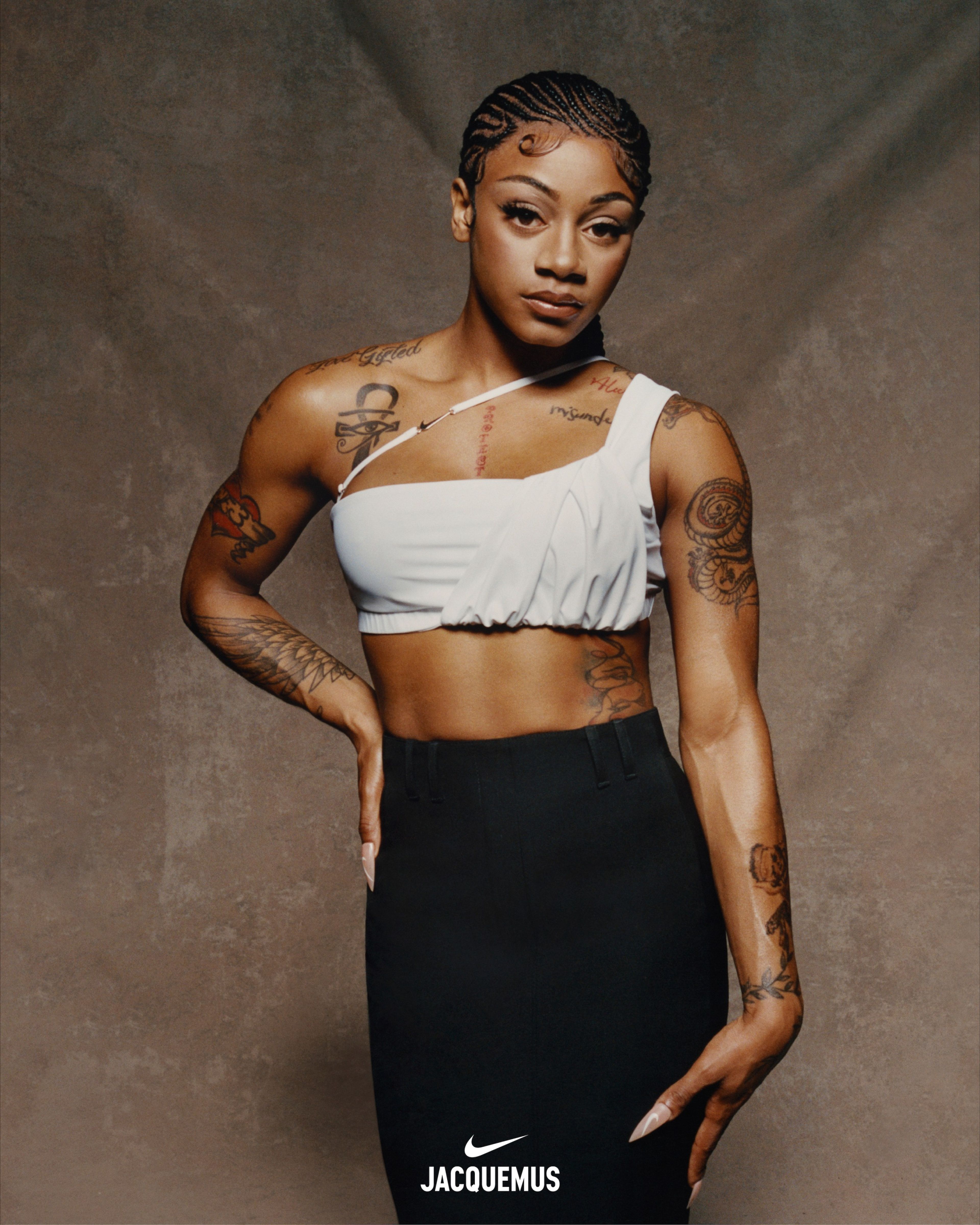 Close up of Sha'Carri Richardson wearing Nike X Jacquemus' slightly ruched white cropped one-shoulder tank and a high-waist maxi-length black skirt. Sha'Carri's hair is in ornate cornrows and we see tattoos on her chest and arms. Sha'Carri looks directly at the camera, not smiling, holding one hand at her hip and the other at her thigh. She stands in front of a beige material background.