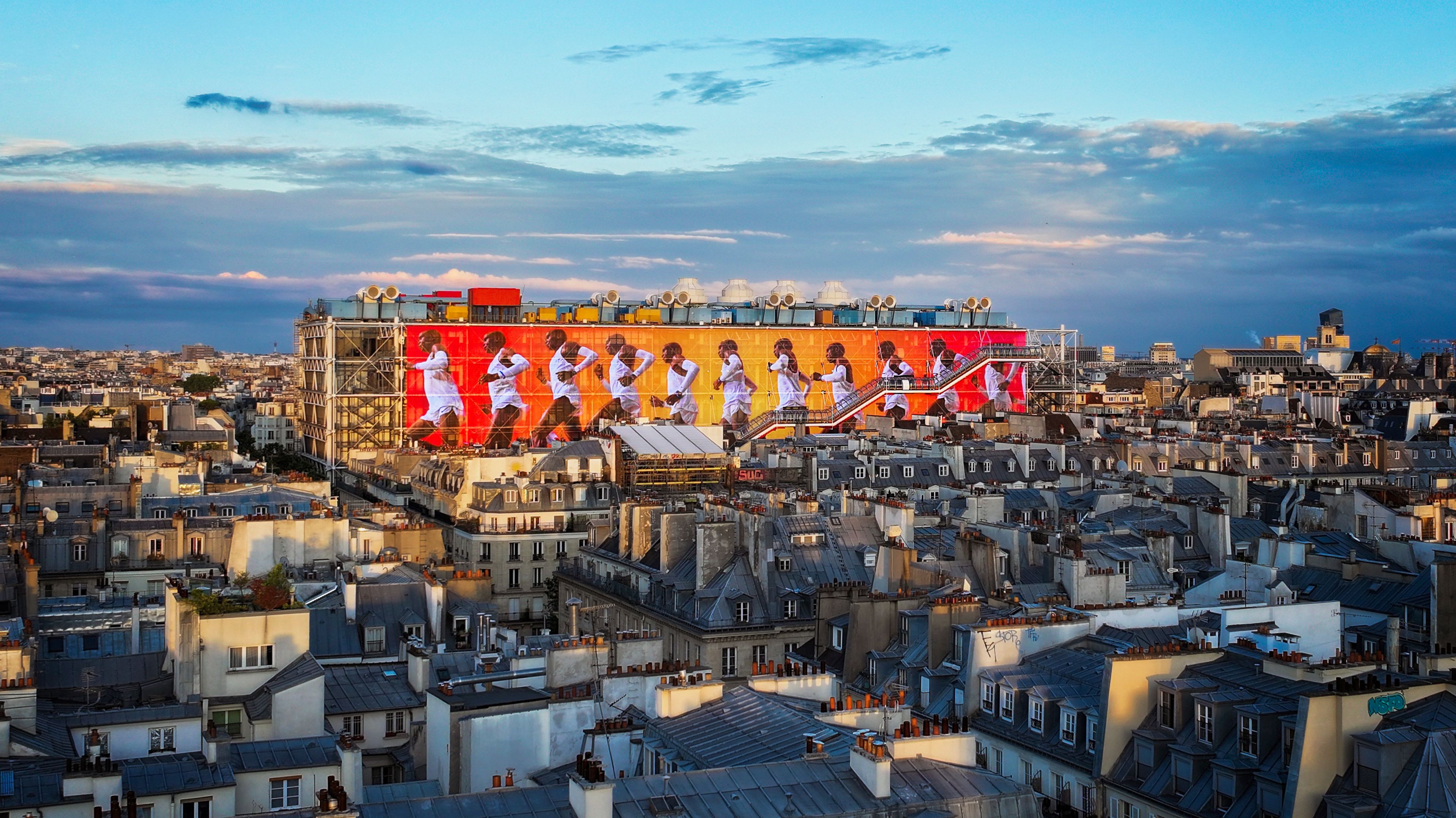 The transformed Nike Au Centre Pompidou’s iconic façade features a canvas that showcases groundbreaking stories of sport. Screen features repeating image of Eliud Kipchoge on bright red and yellow colors, which pops out against the cityscape and a cloudy blue Paris sky.