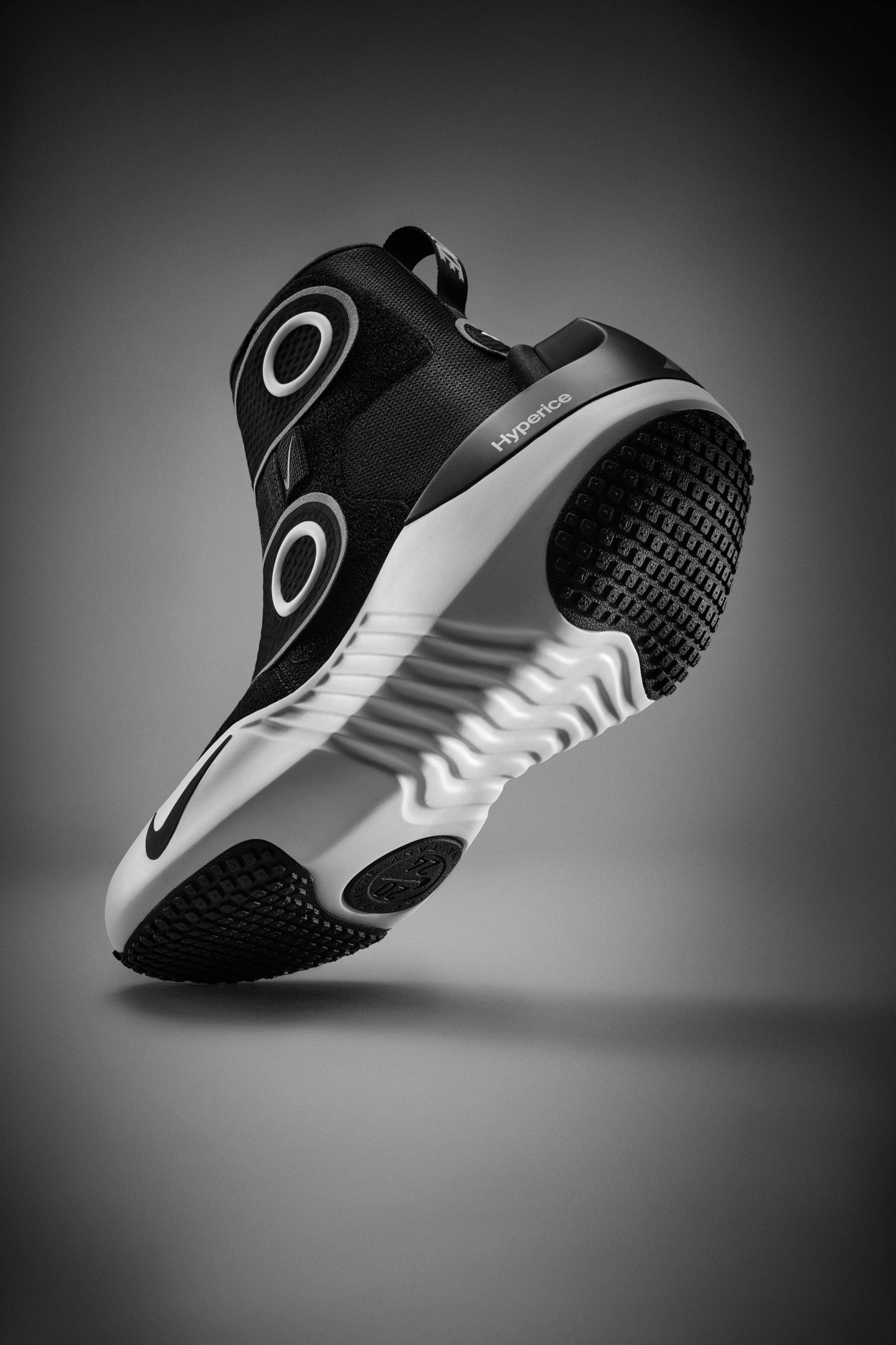 Underside view of the Nike x Hyperice boot, which is a high-top boot with a black upper, white midsole, and black and white sole. We see black waffle pattern pods at the toe and heel of the sole. The boot's toe sits on a grey floor, while the heel floats above, casting a shadow underneath.