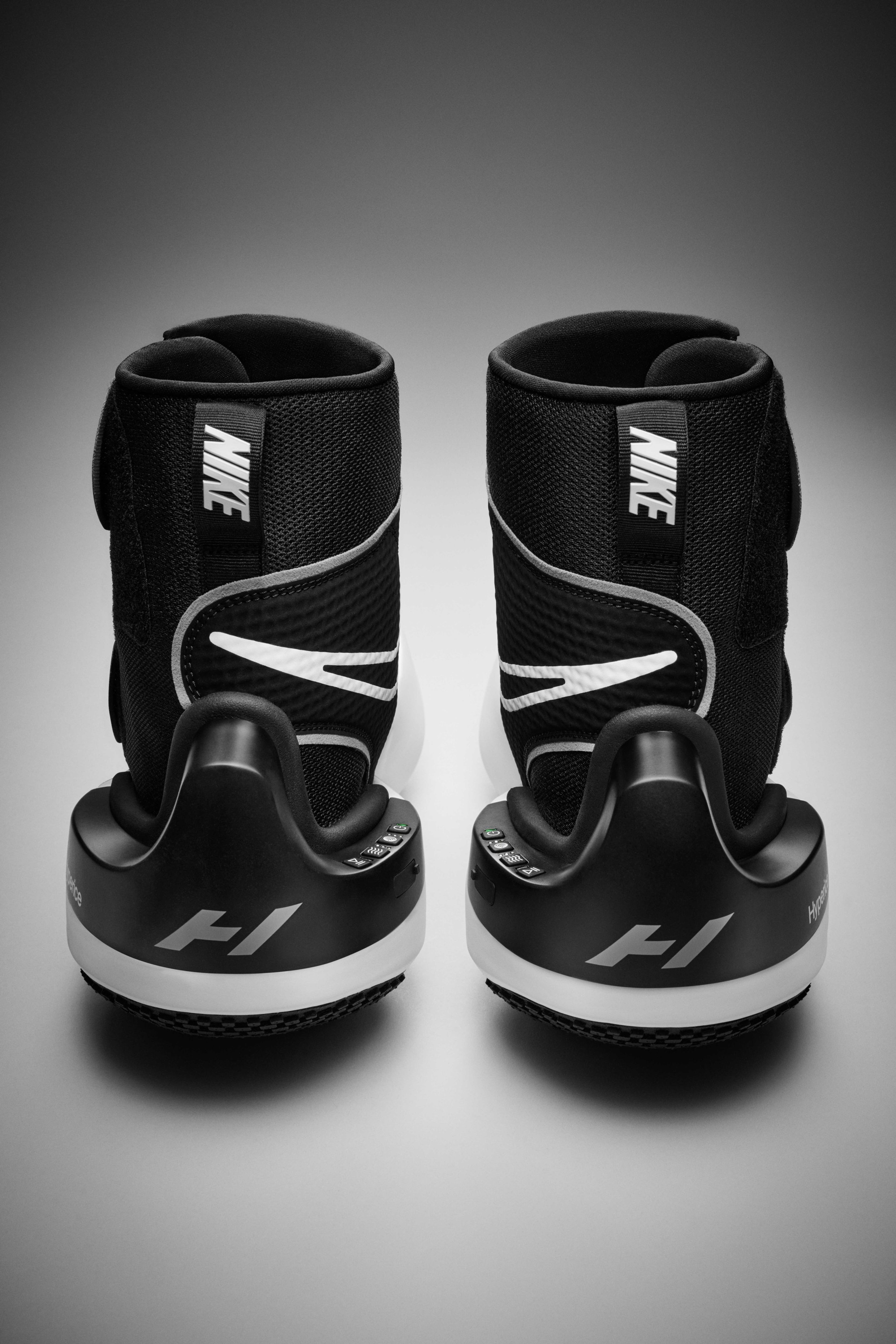 Back view of a pair of Nike x Hyperice boots, detailing the control units on each shoe. Black boots sit on a grey surface.