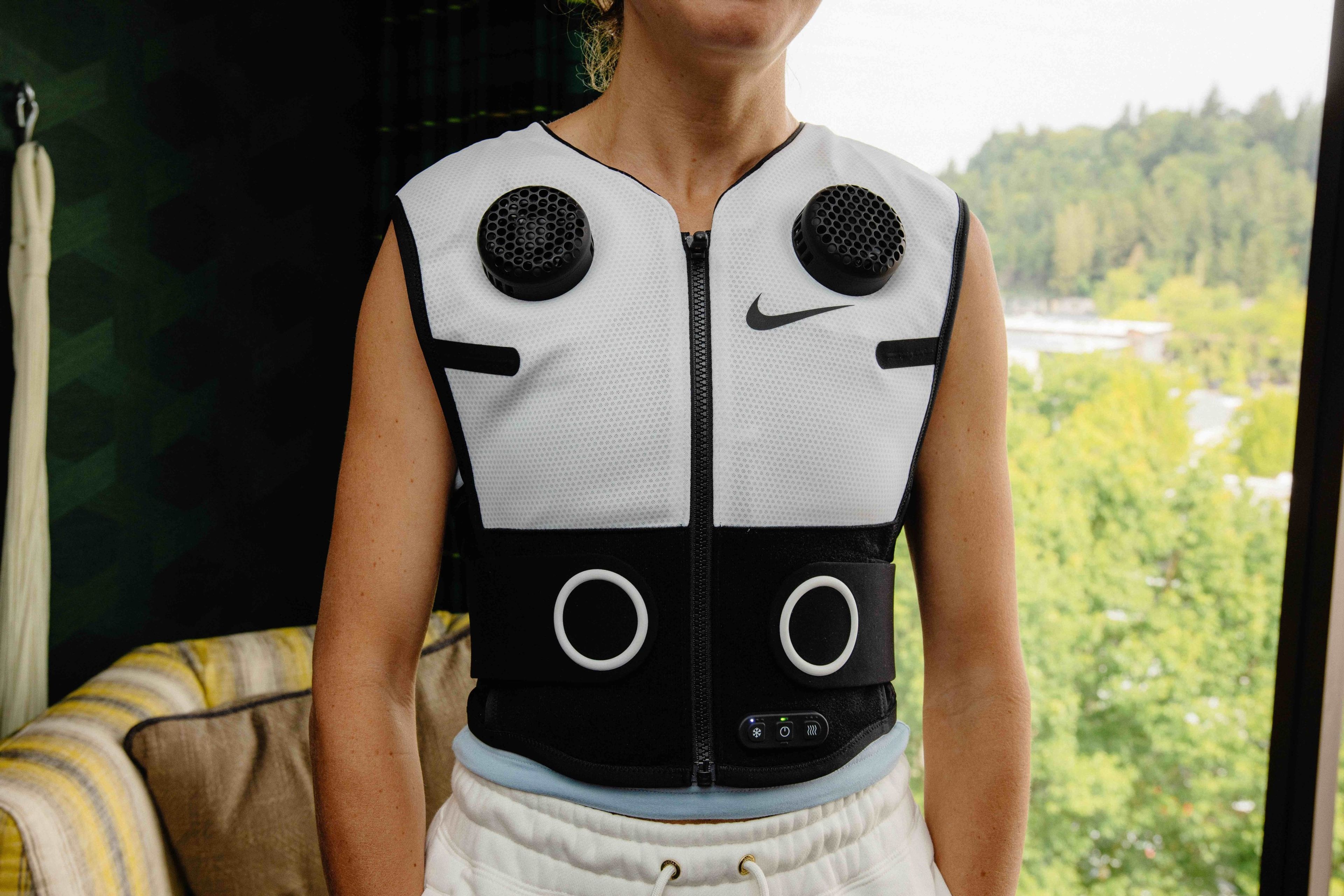 Front view of an athlete wearing the white Nike x Hyperice vest. We see the torso of the athlete wearing the vest with white fleece pants while standing in front a window with trees in the background.