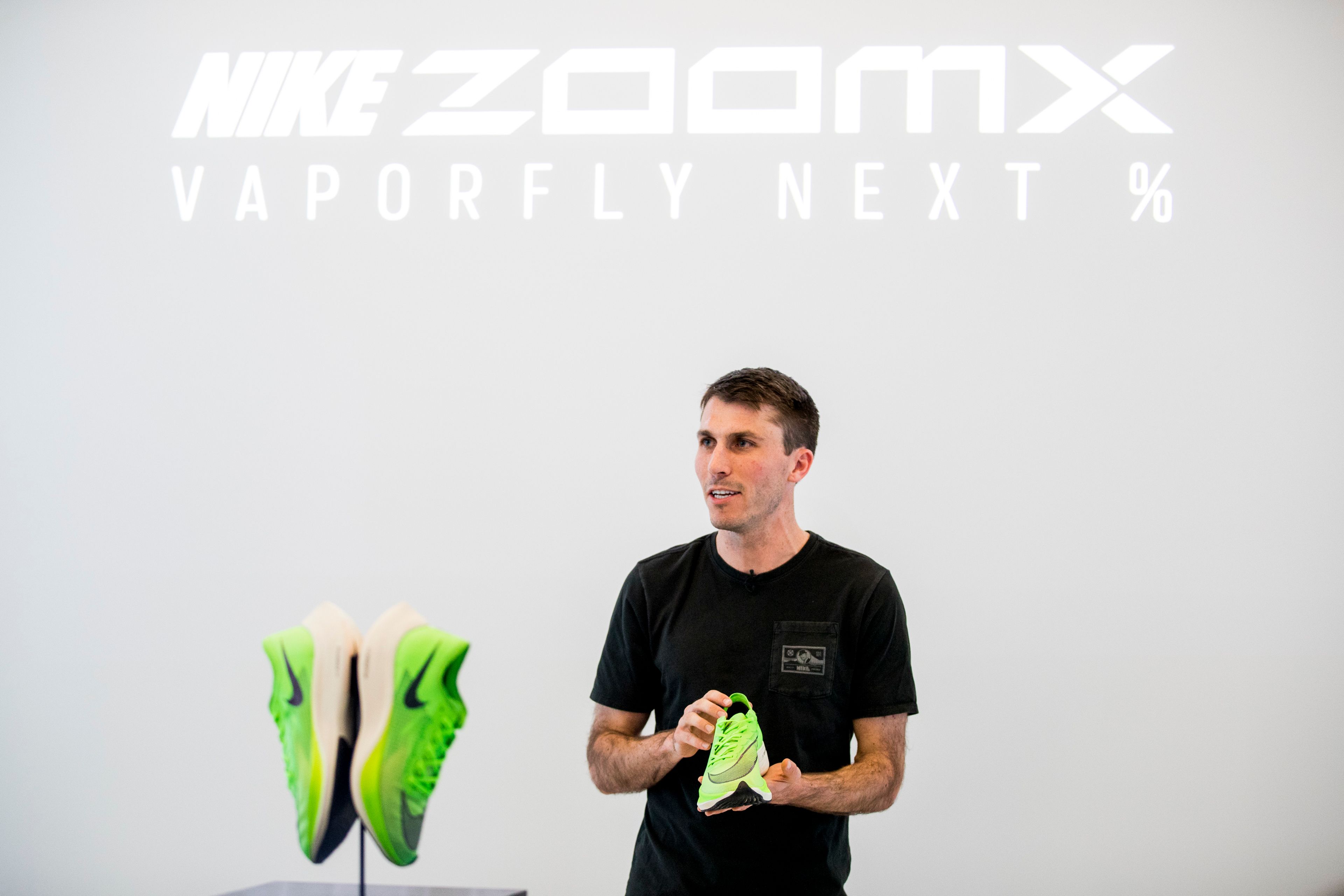 Elliott Heath in black T-shirt holding a neon green Nike ZoomX Vaporfly Next% shoe at a brand event, with a second pair displayed beside him.