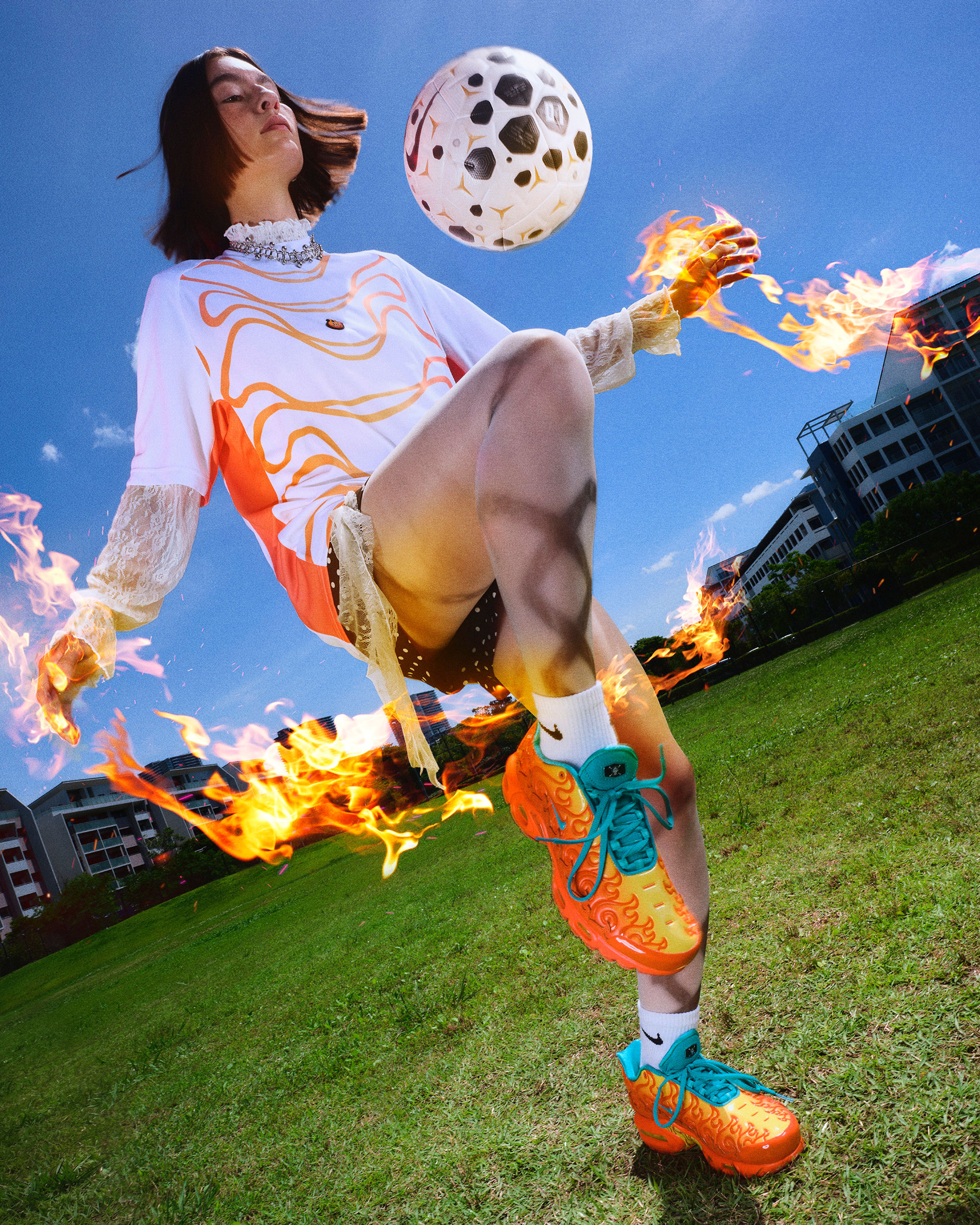 A woman in orange and yellow Nike sneakers with teal laces performs a soccer kick with flames trailing from her hands and feet against a bright blue sky.