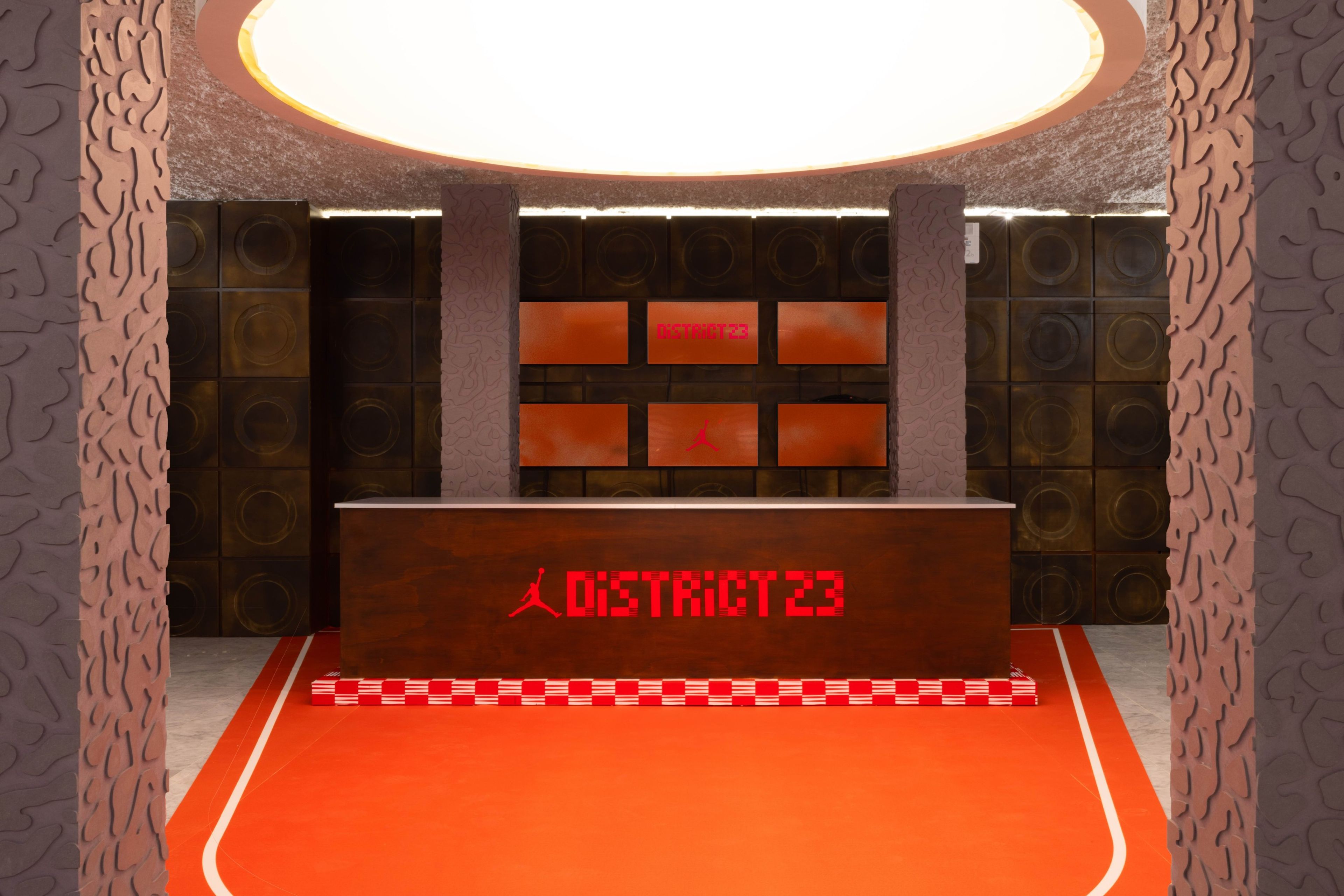 A photo of the District 23 main entrance area, featuring a front desk, orange flooring, a circular overhead light, and digital displays behind the desk.