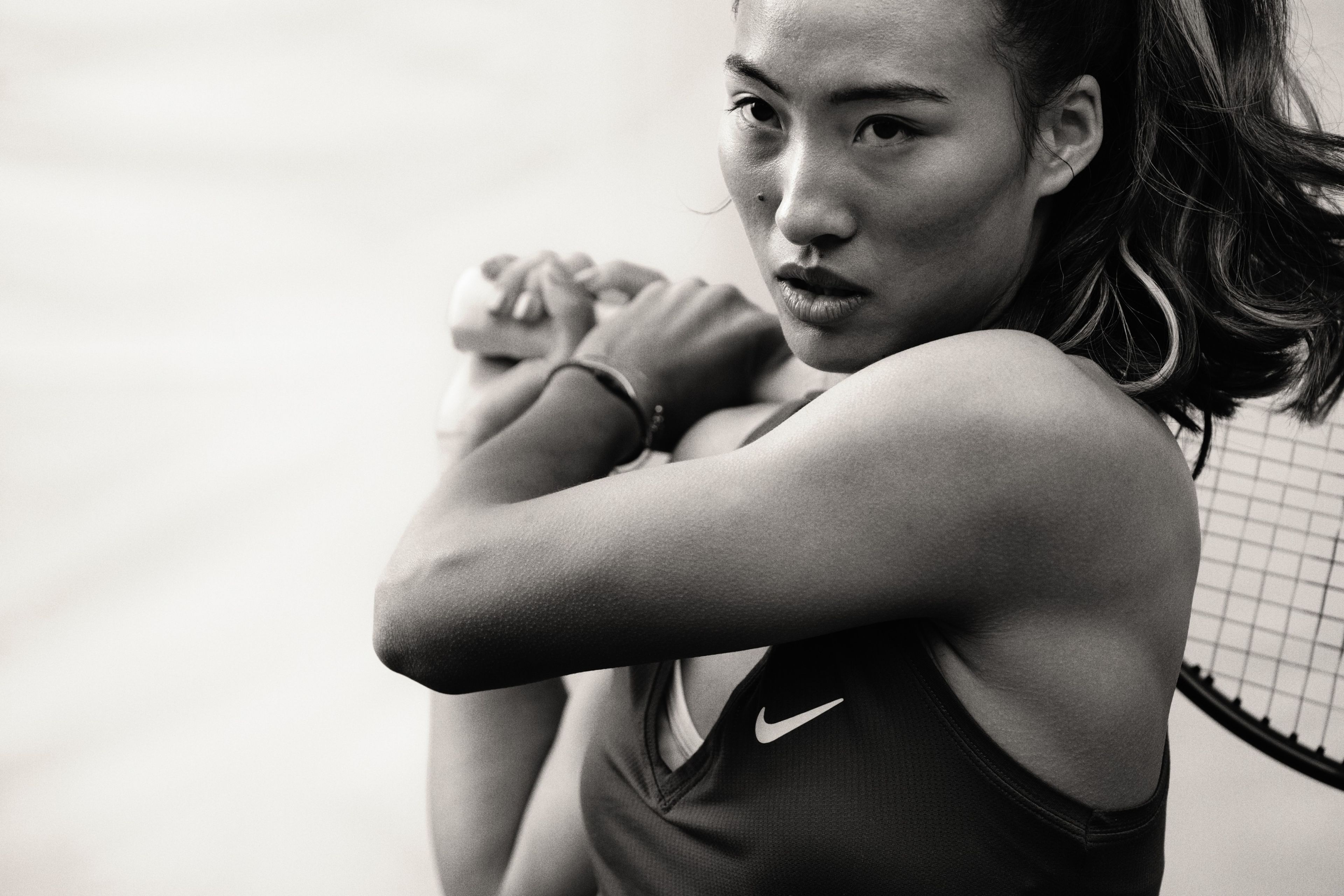 Black and white close up photo of Qinwen Zheng holding a tennis racquet over her shoulder with both hands. She wears a black NikeCourt Victory tank with a white Swoosh.