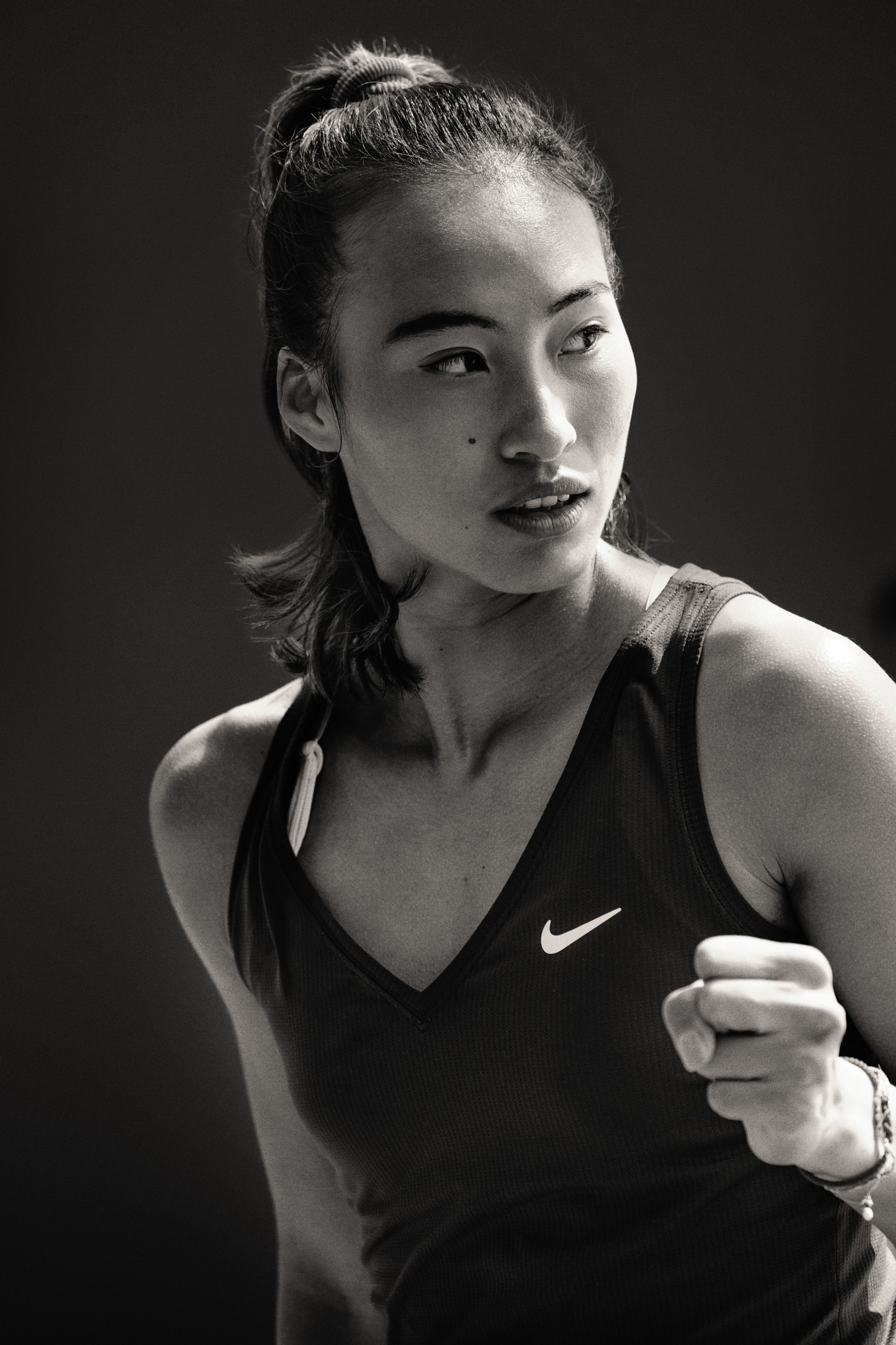 Black and white close up photo of Qinwen Zheng, wearing the NikeCourt Victory Tennis Tank in black. She looks across the tennis court and holds her hand in a fist.