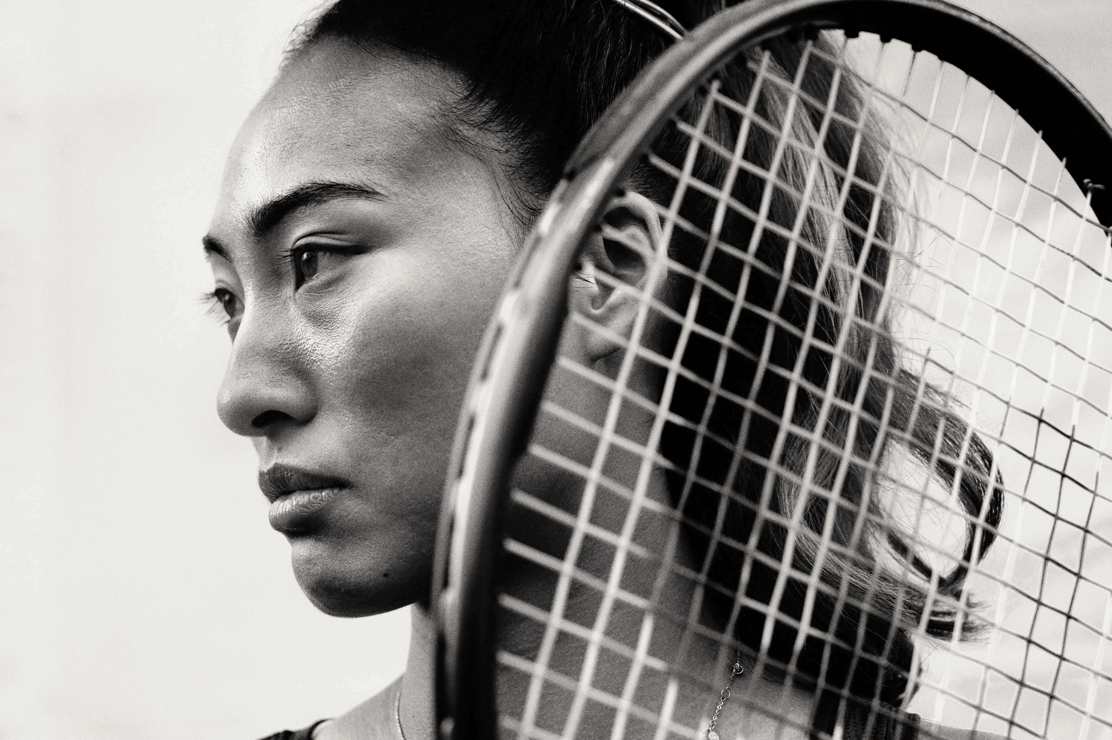 Black and white close up of Qinwen Zheng holding her tennis racquet near her face. Her hair is pulled up into a high ponytail and she looks to the left.