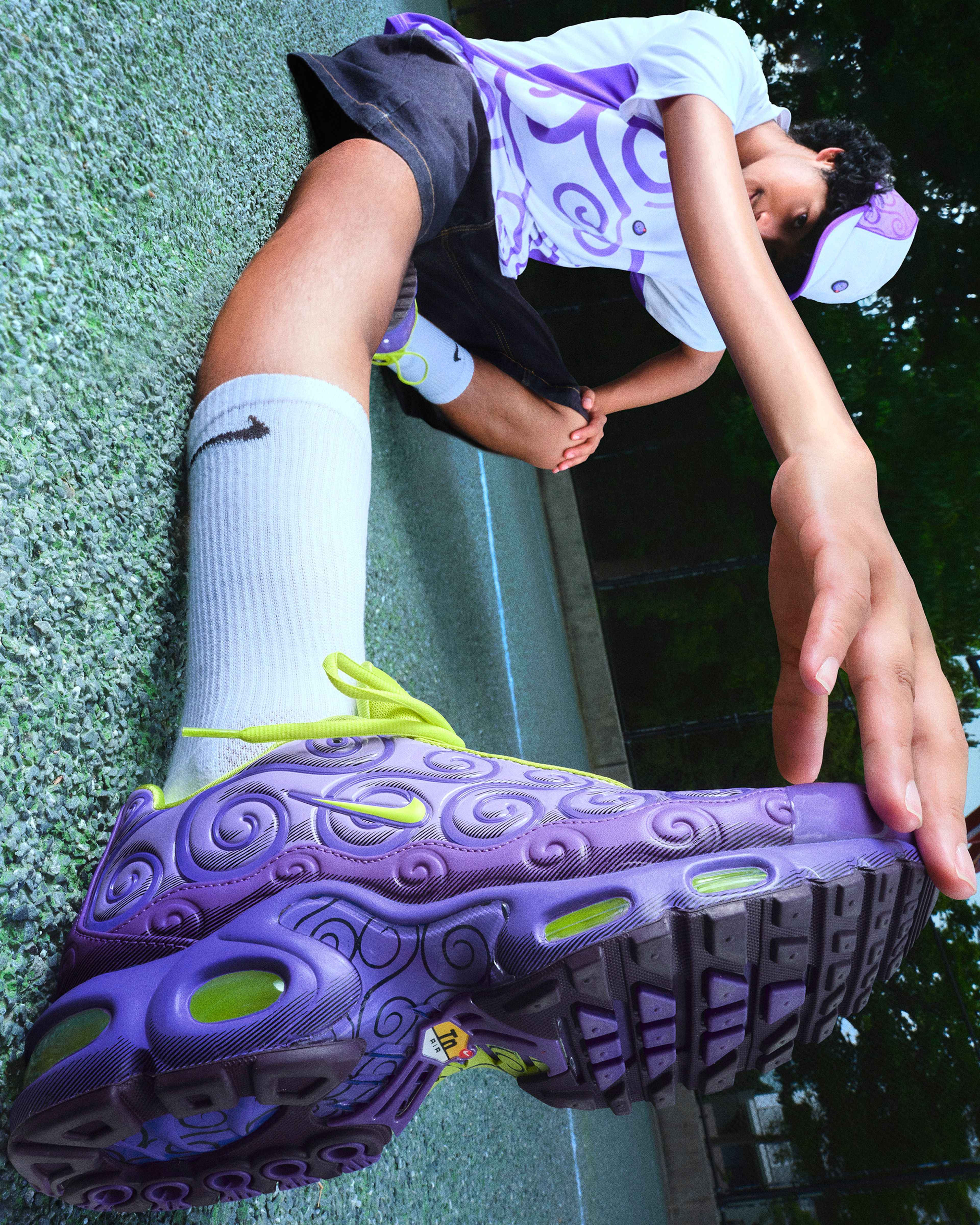 A person wearing a purple and white patterned shirt and matching cap reaches toward a purple Nike sneaker with neon green laces on an outdoor court.