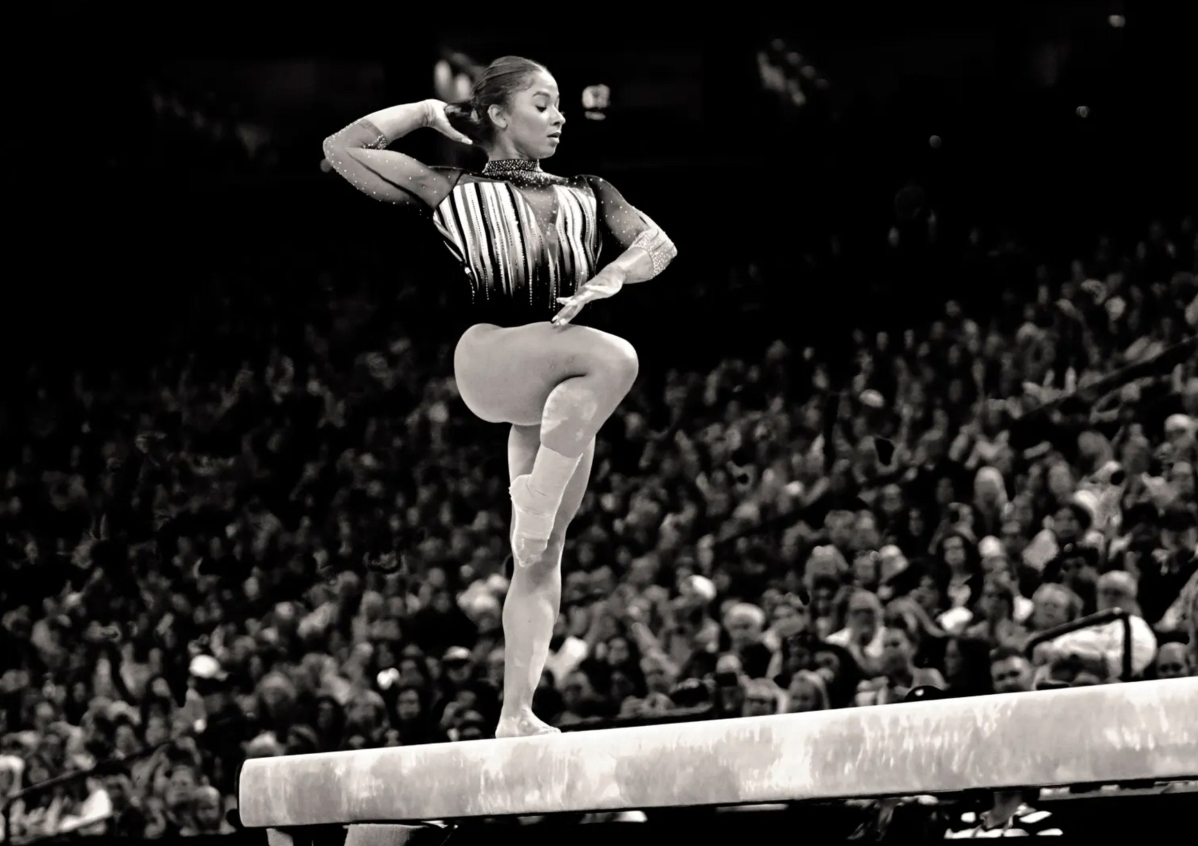 A black and white photograph of Jordan Chiles of Team USA Women’s Gymnastics taken while she's in competition on the balance beam. With a full crowd behind her, she lifts her knee and looks to her right.
