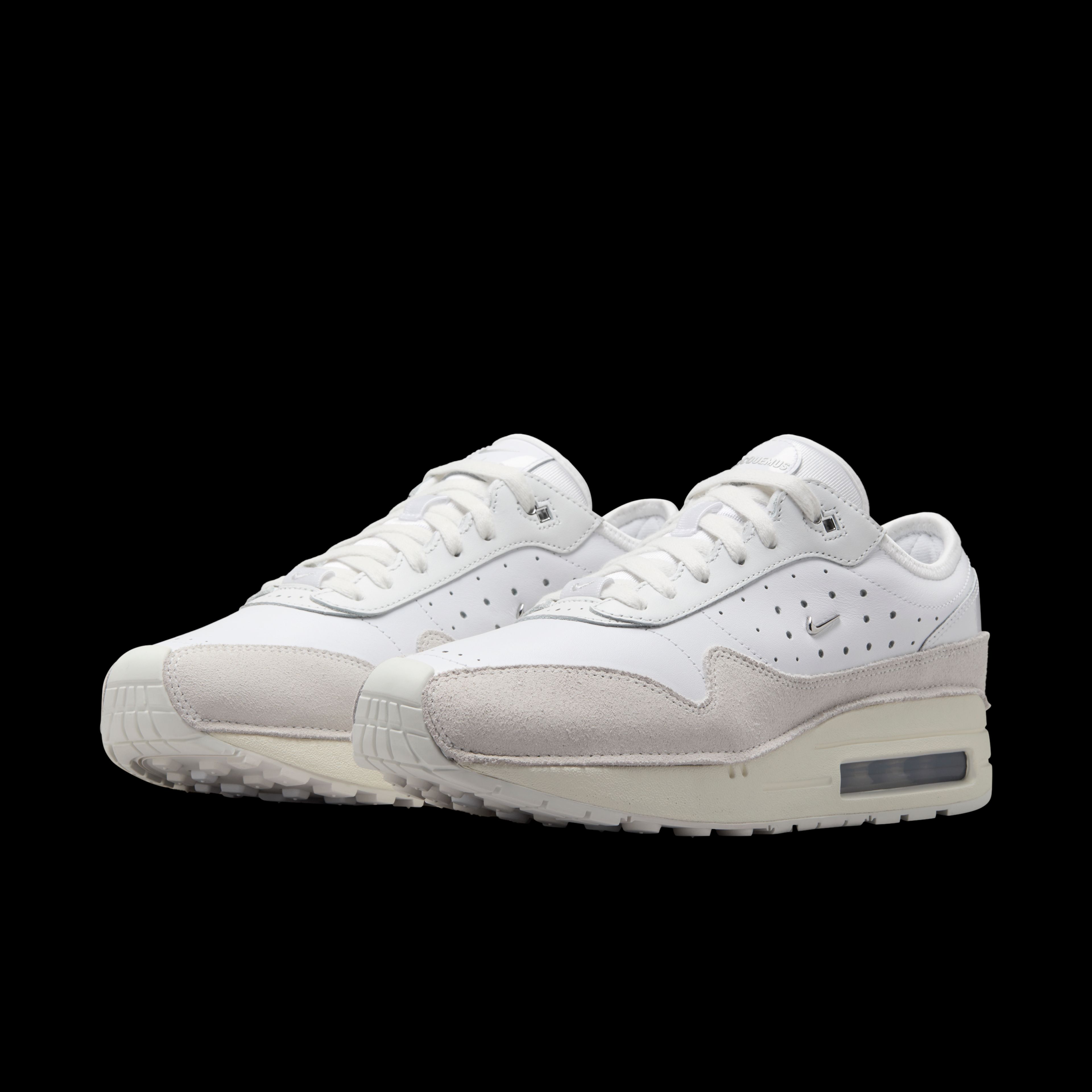 Front angled view of a pair of Nike x Jacquemus Air Max 1 sneakers. The shoes have a white upper with sail suede details, a white midsole, and white sole. There is a small metallic silver Swoosh on the side of the shoe. The pair floats on a black background.