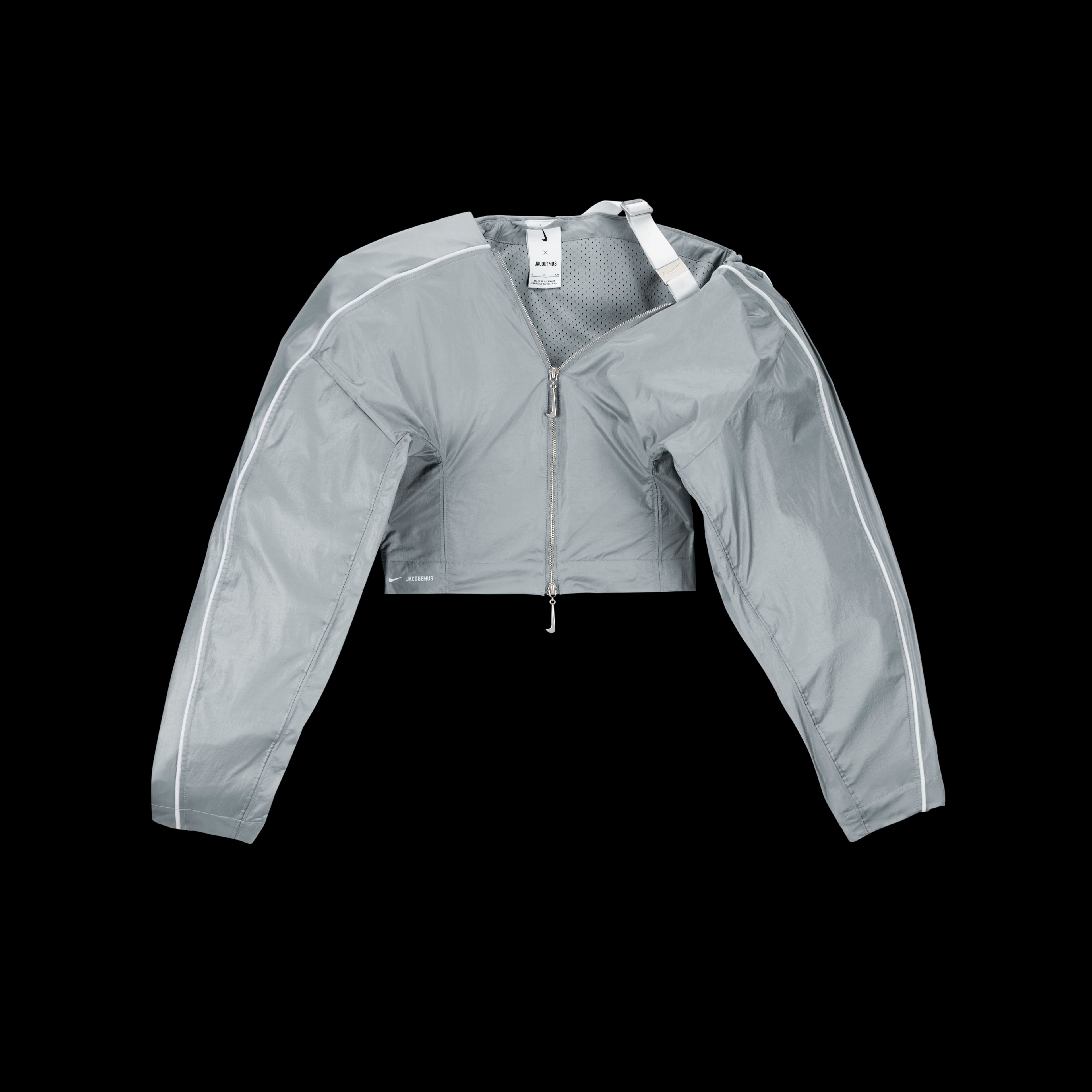 Overhead view of the Nike x Jacquemus Women's Track Jacket in particle grey with white pinstripes across the shoulder and down the sleeve. One sleeve is oversized and attached to the back of the jacket with white webbing. The jacket zipper pull is grey in the shape of a Nike Swoosh. The piece floats on a black background.