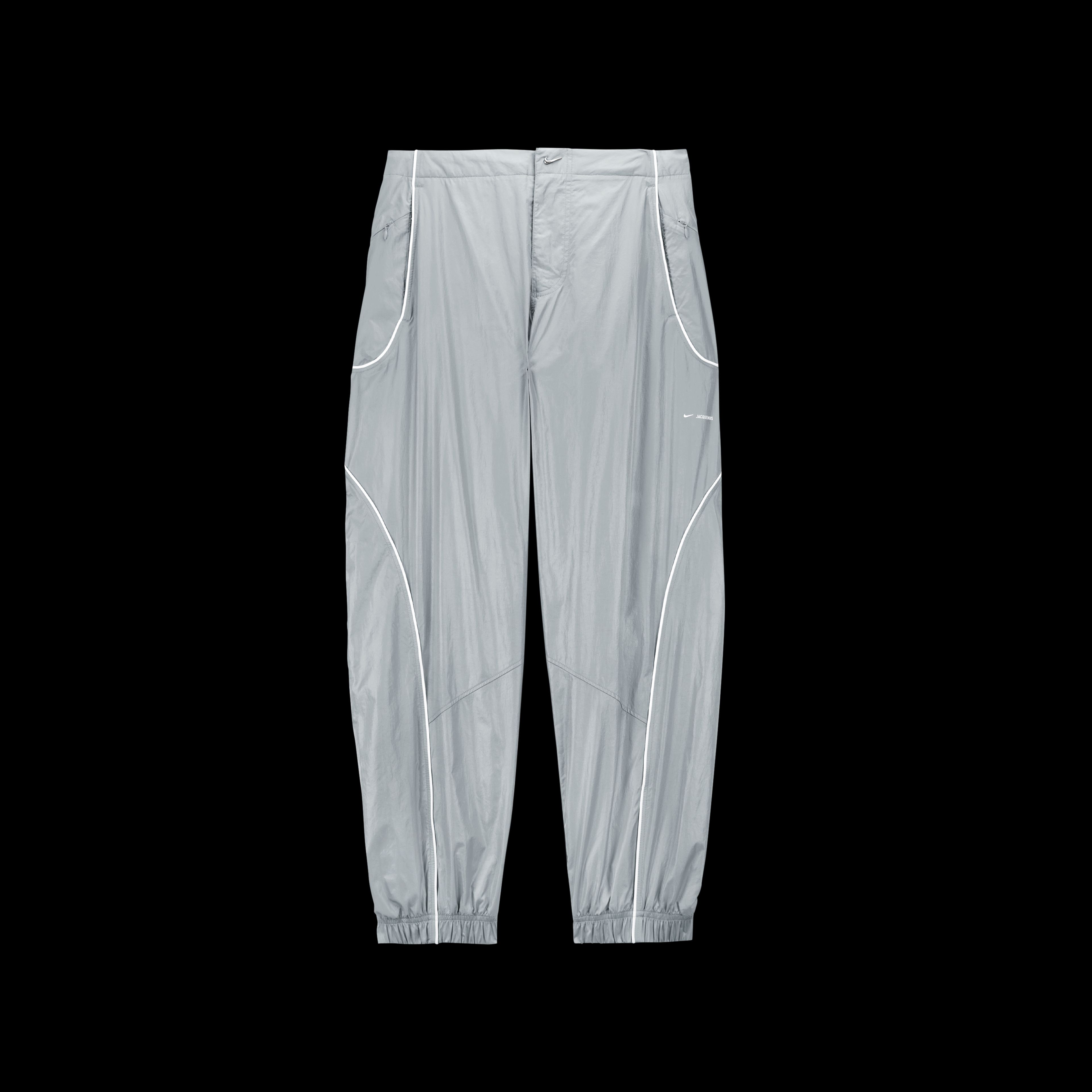 Overhead view of the Nike x Jacquemus Track Pant in particle grey with curved white pinstripes at the pocket and down the leg. There is a small shiny silver swoosh as a closure on the waist, and elastic cuffs at the ankle. The pant floats on a black background.