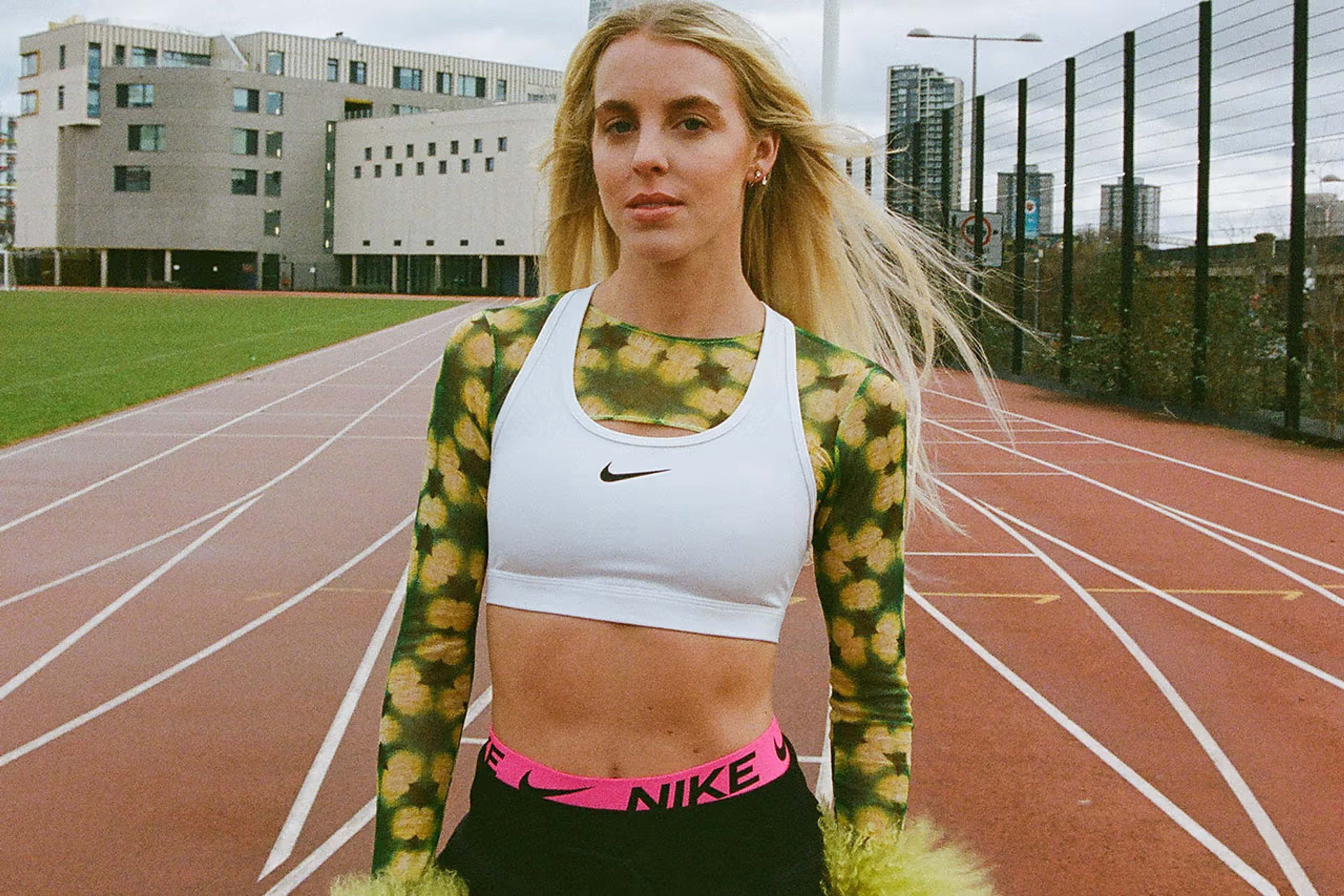 Close up of Keely Hodgkinson standing on a Track. She wears a white Swoosh bra layered over a green and black crop top, and black pants with the pink band of Nike Pro shorts visible. A cityscape is visible in the background.