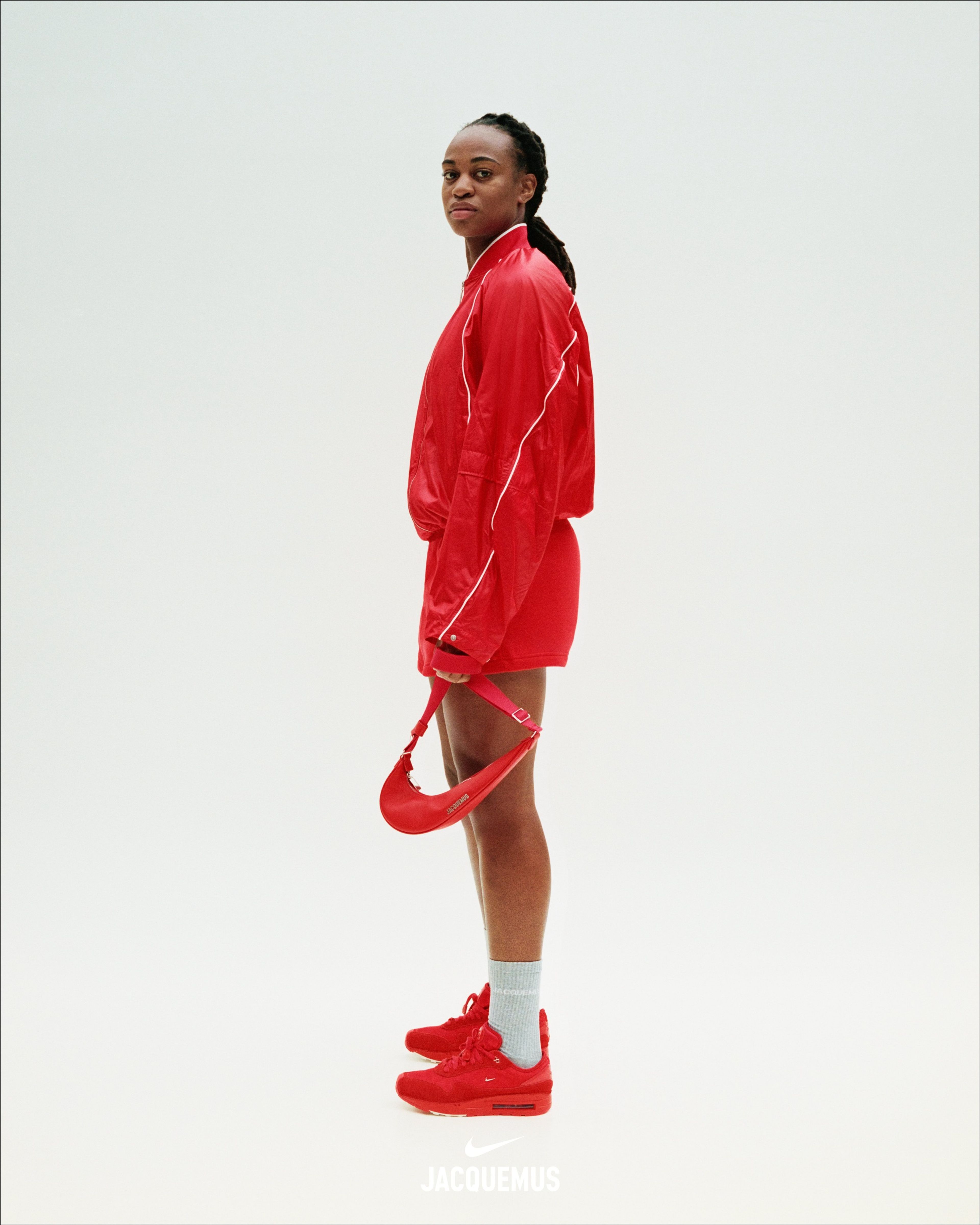 Side view of Marie-Antoinette Katoto wearing a red Nike x Jacquemus Track Jacket, with red shorts, light grey socks, and the Nike x Jacquemus Women's Air Max 1 '86 Big Bubble in Mystic Red. Marie-antoinette faces left as she stands on a white background, looking at the camera and holding a red Nike x Jacquemus Le Swoosh Crossbody Bag in her hand.