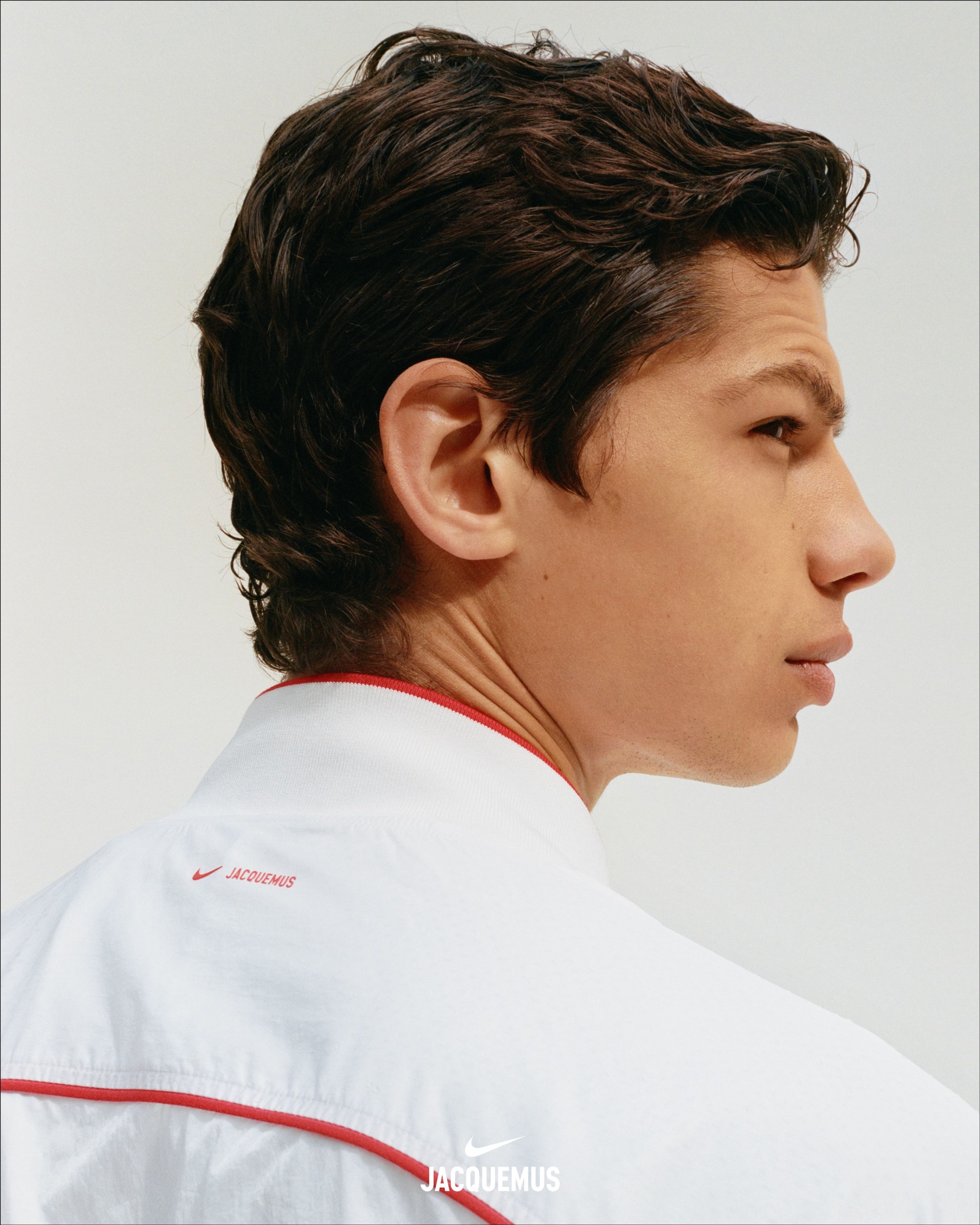 Close up head shot of a male athlete wearing the Nike x Jacquemus Track Jacket in white with university red piping at neckline and across back. We see a red Nike Swoosh and Jacquemas logos. The man stands against a white background and looks to the right.