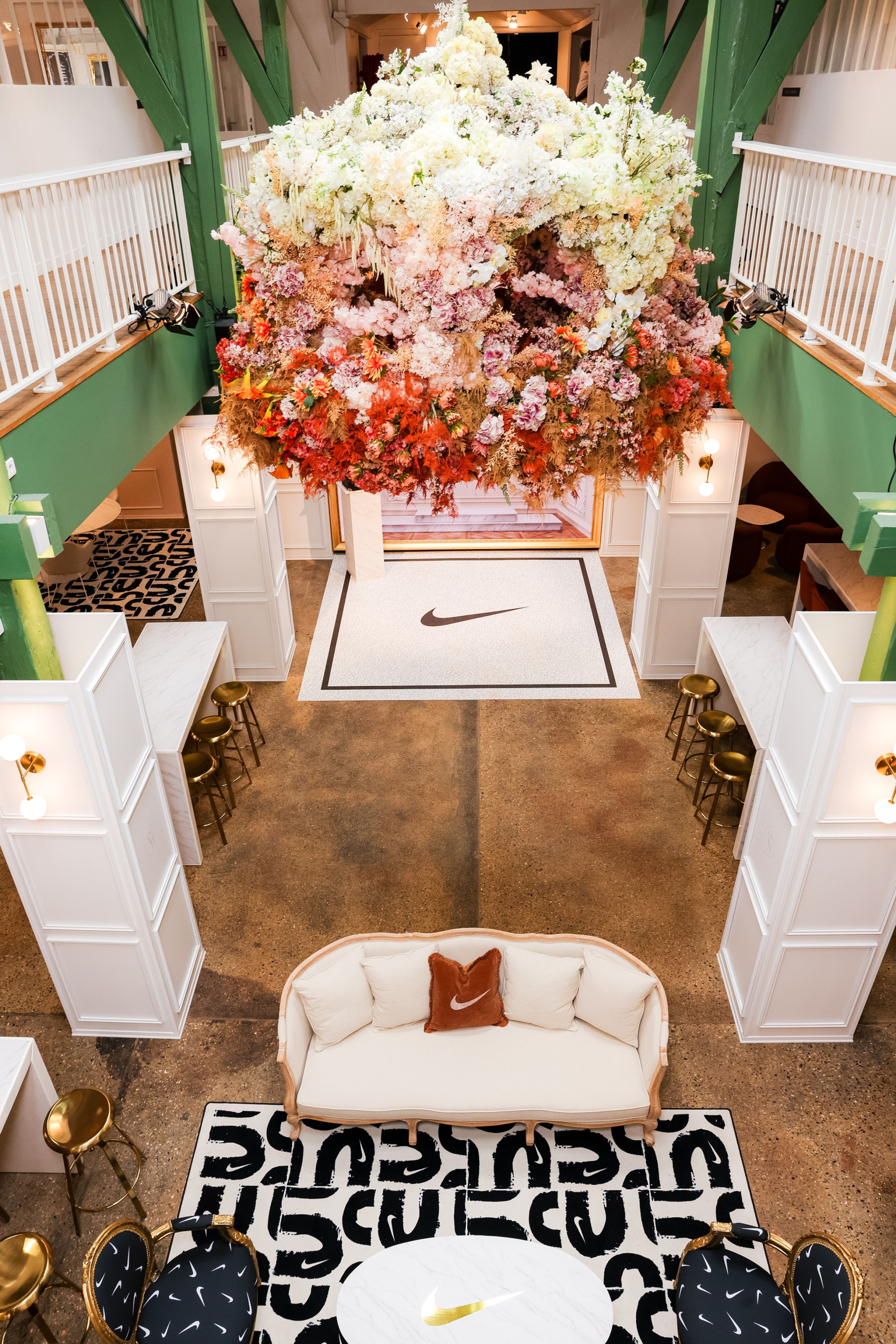 Overhead view into the entryway of the Nike Athlete House in Paris, France. A dramatic flower display hangs above a lounge space with green walls. 2 chairs are upholstered with black fabric with white Swooshes. A white sofa holds a rust colored pillow with a white Swoosh. There is white cabinetry and high tables with barstools flanking the room.