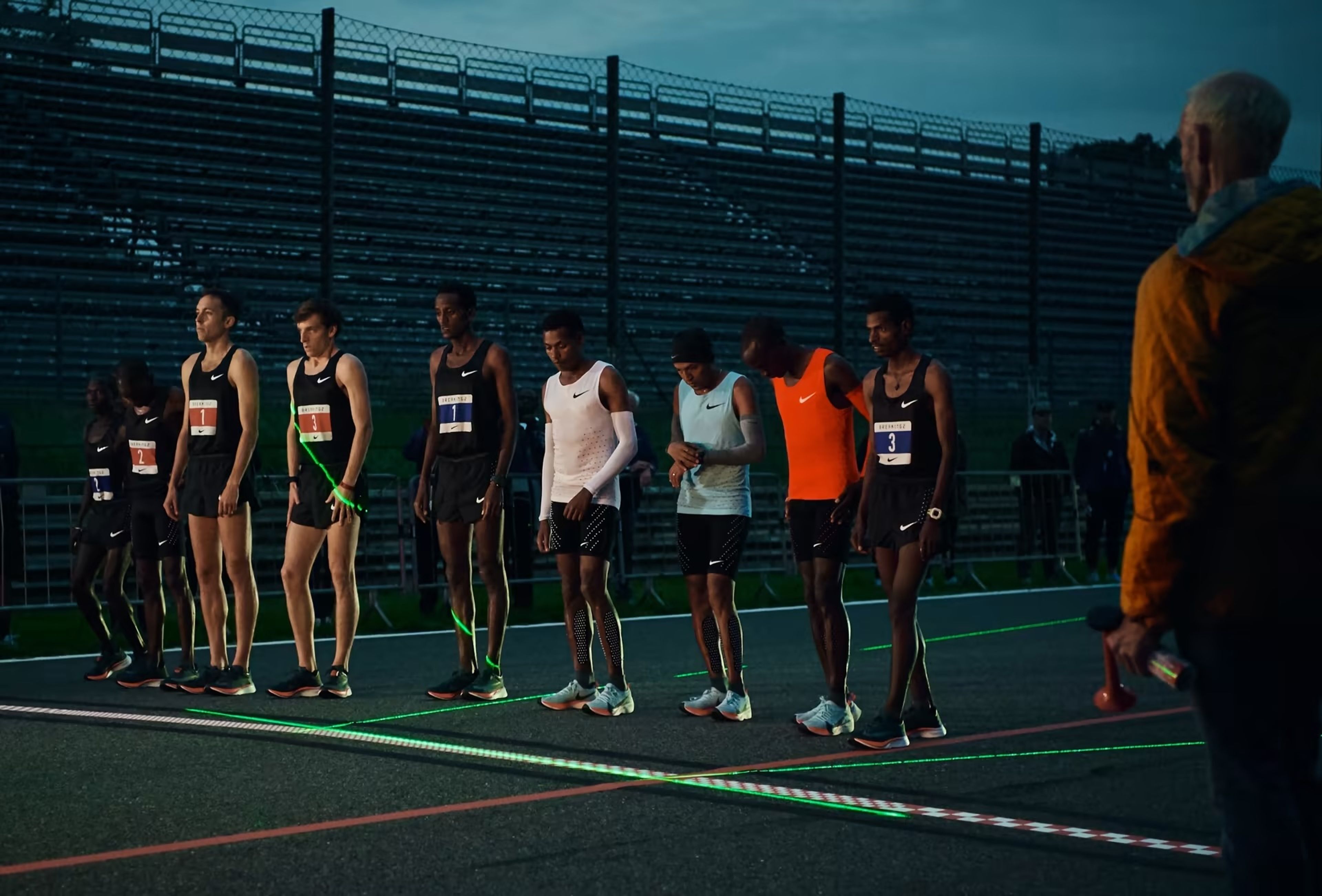 Runners line up for the Breaking2 event at the Monza Circuit track at dusk, illuminated by green laser lines, with an official holding a starting gun nearby.