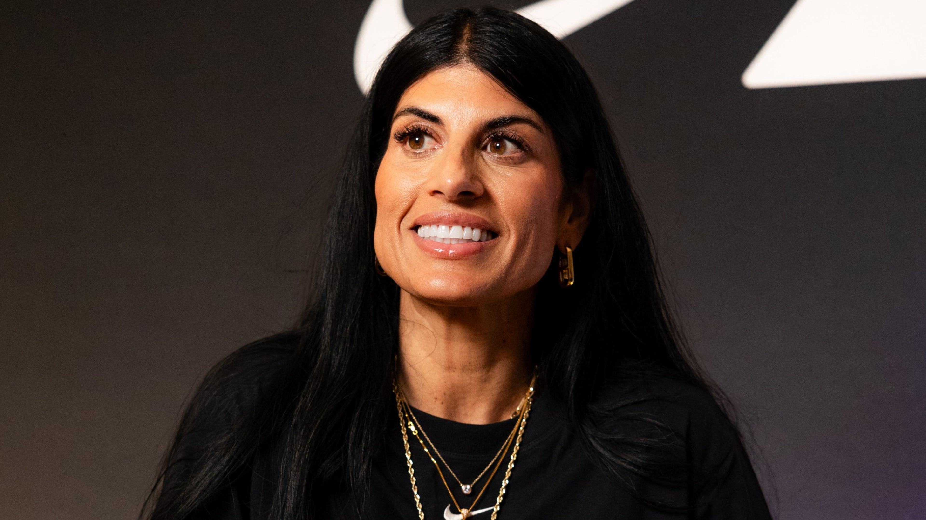 Close up of Nike track and field coach, Diljeet Taylor, wearing a black Nike Sportswear Chill Knit dress, with layered gold jewelry. She sits in a white chair, against a dark background, looking slightly to the left.