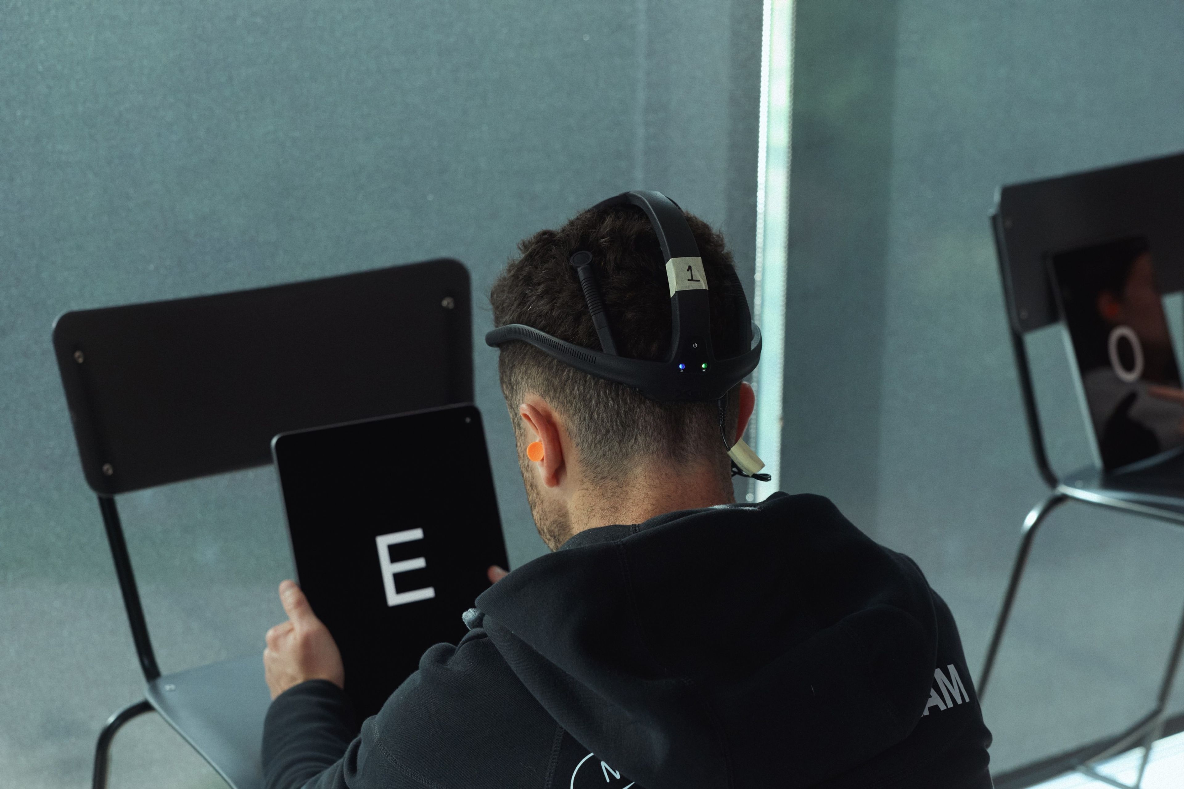 An adult wearing a black hoodie and VR headset with sensors holds a tablet displaying the letter E during a cognitive testing session.