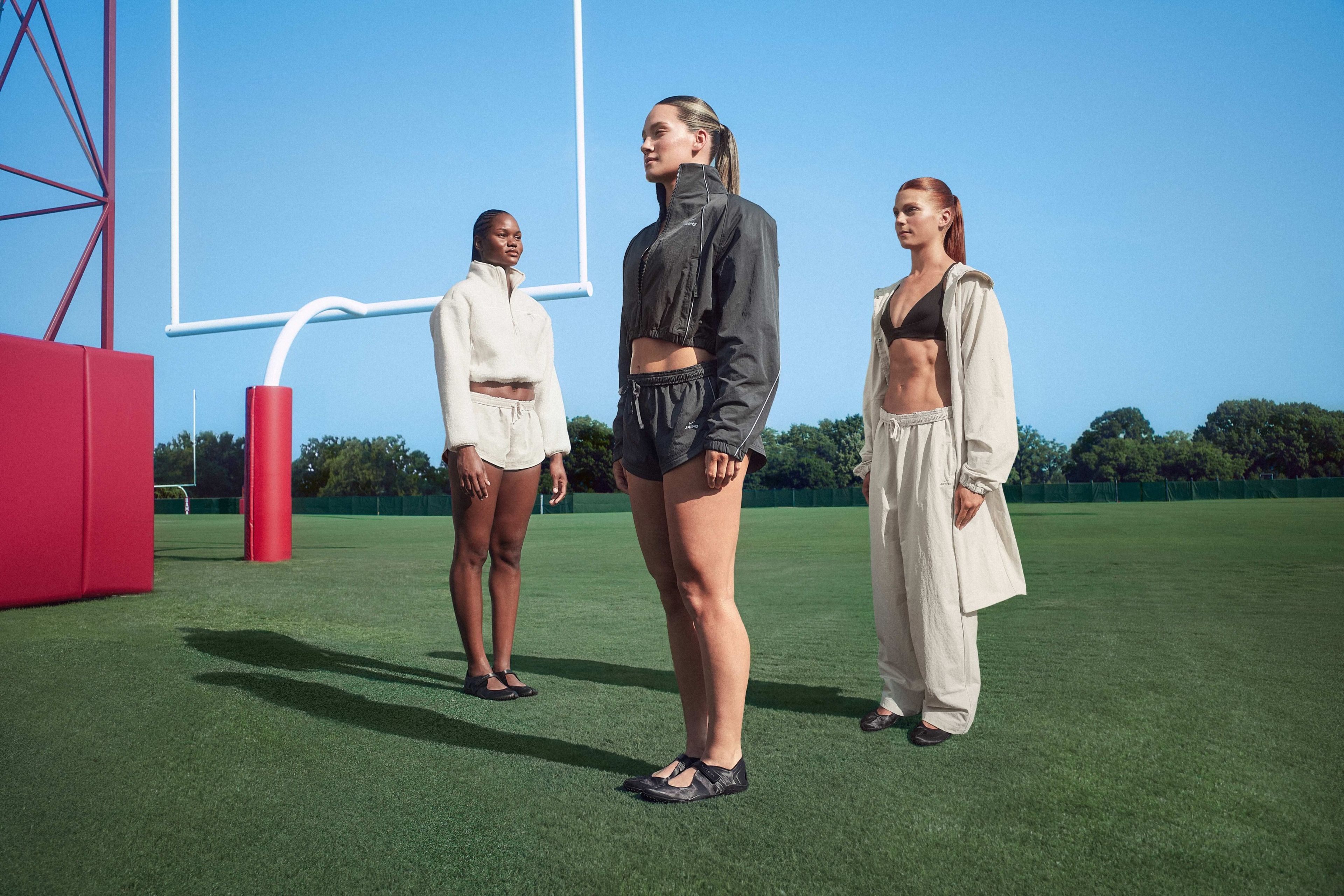 Three women in NikeSKIMS apparel stand on a football field turf near a goal post, wearing neutral-toned crop jackets, shorts, sports bra, and an oversized trench coat with NikeSKIMS footwear