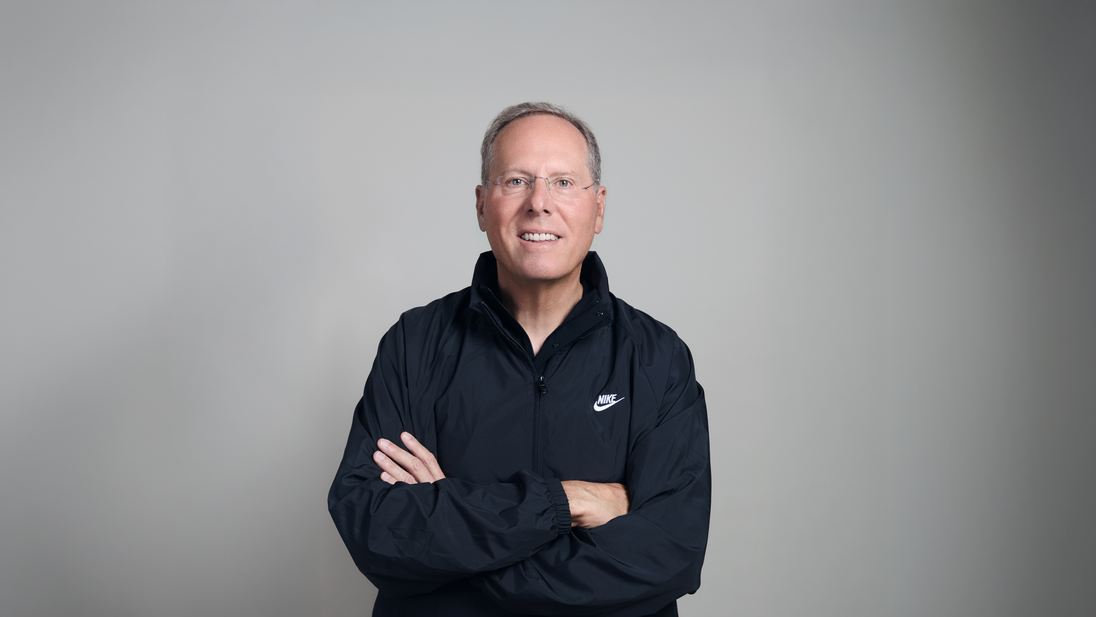 Dave Denton, Executive Vice President and Chief Financial Officer, NIKE, Inc., poses with his arms crossed in front of a gray background. He's wearing a black zip-up Nike jacket.