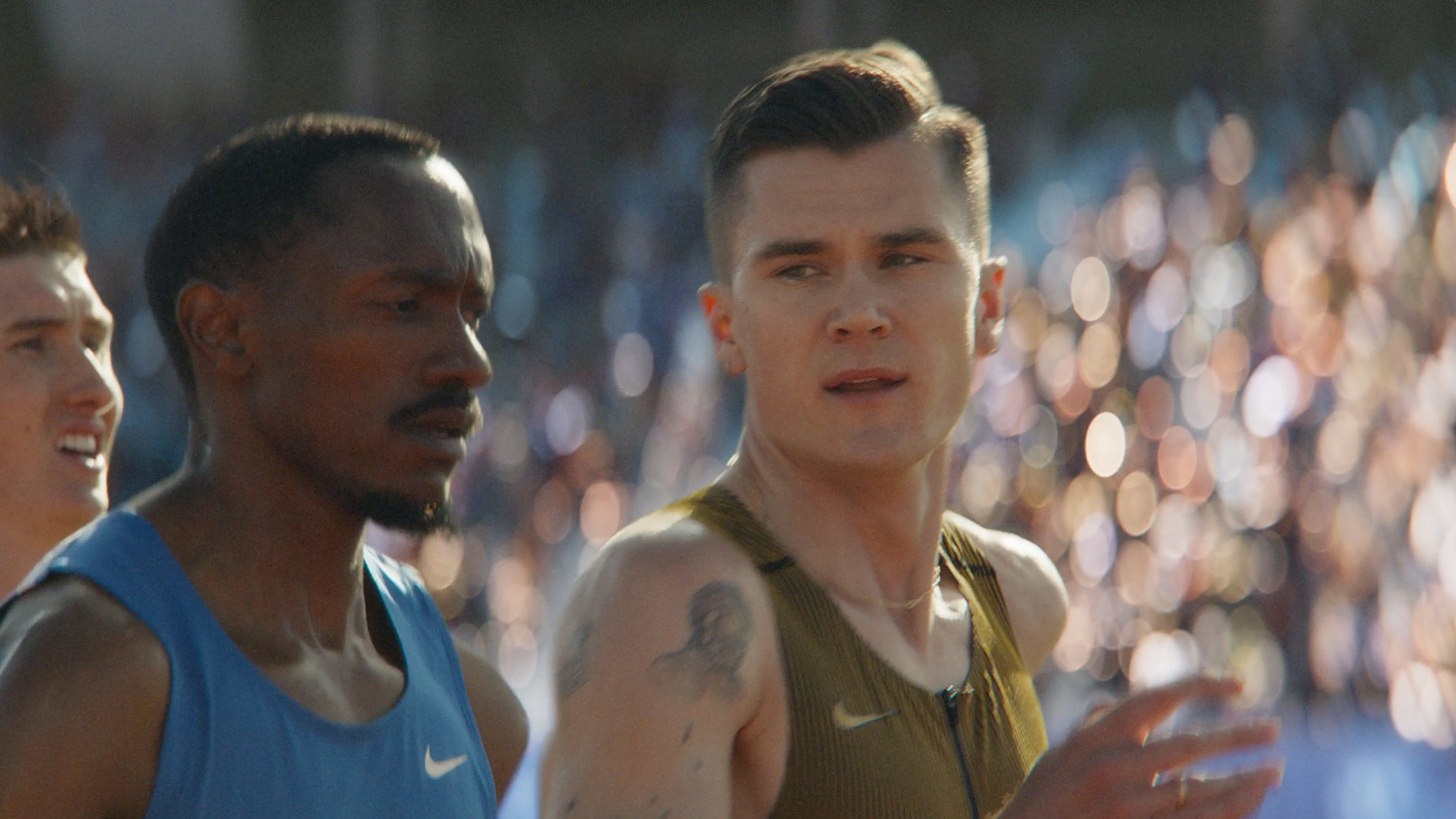 Close up of Jacob Ingebrigtsen at a track event, alongside another competitor. Jacob wears a gold Nike running singlet and there is a crowd in the background.