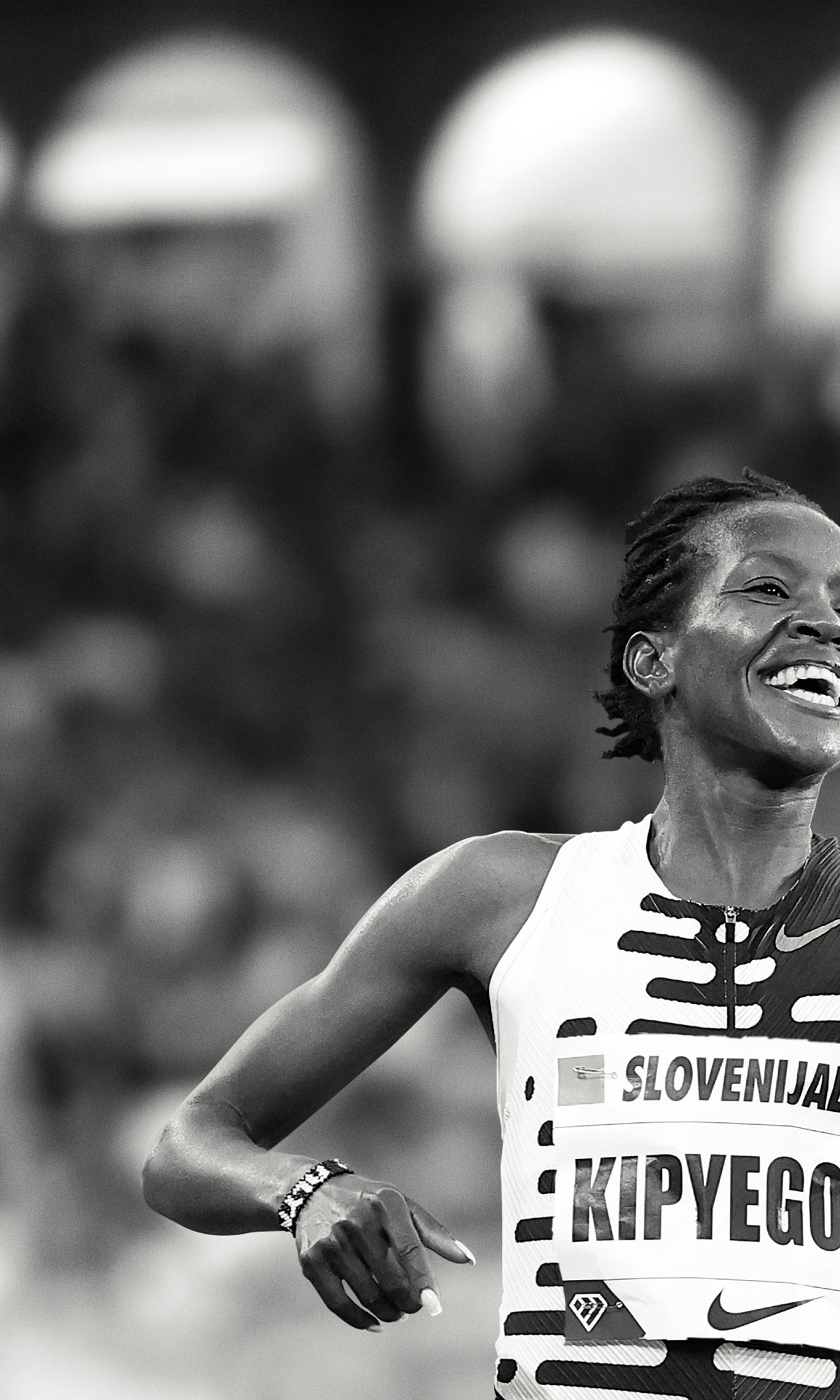 Black and white photo of Faith Kipyegon with a celebration smile and pointing a finger forward. She wears a patterned Nike running top, and there is a crowd behind her.