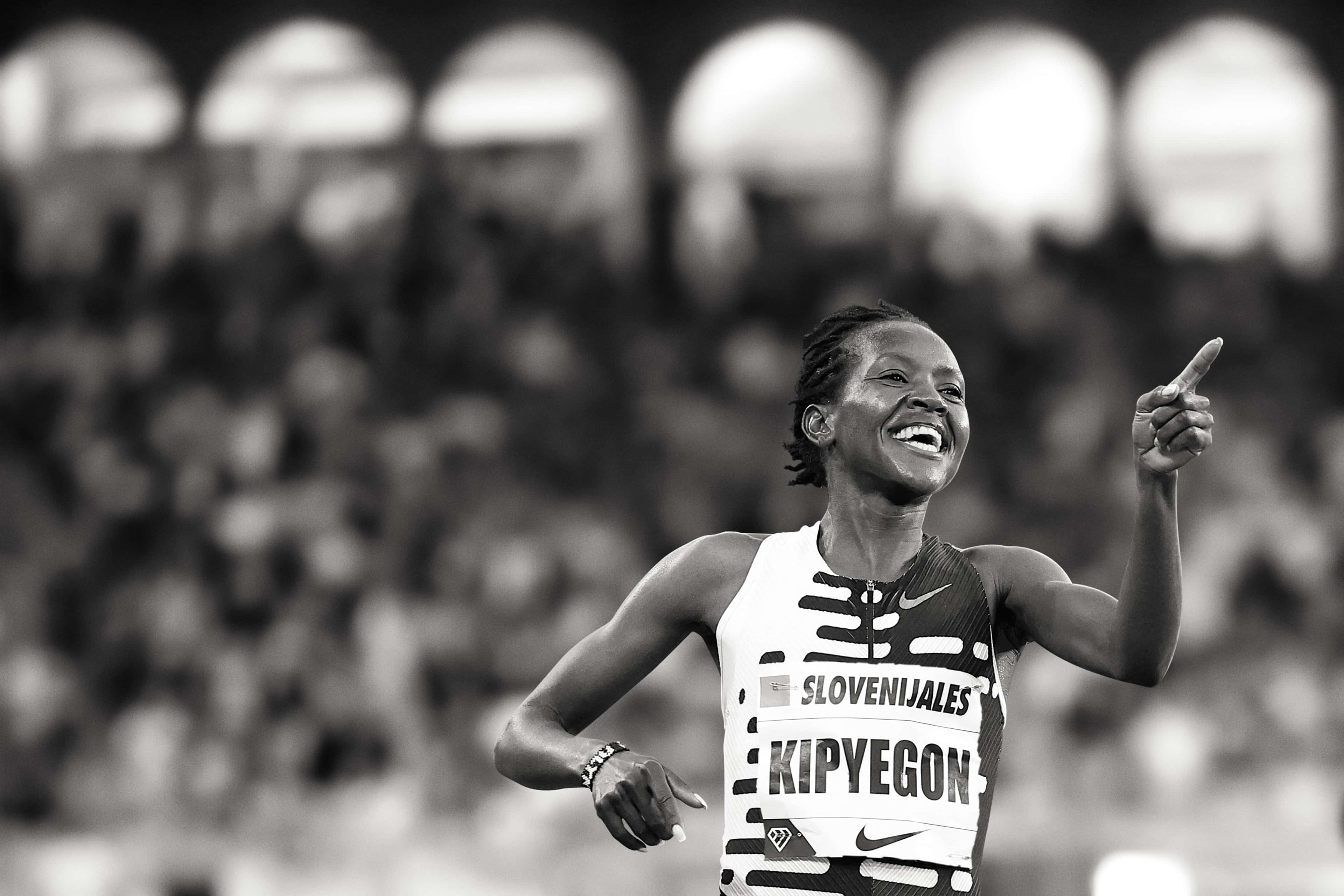 Black and white photo of Faith Kipyegon with a celebration smile and pointing a finger forward. She wears a patterned Nike running top, and there is a crowd behind her.
