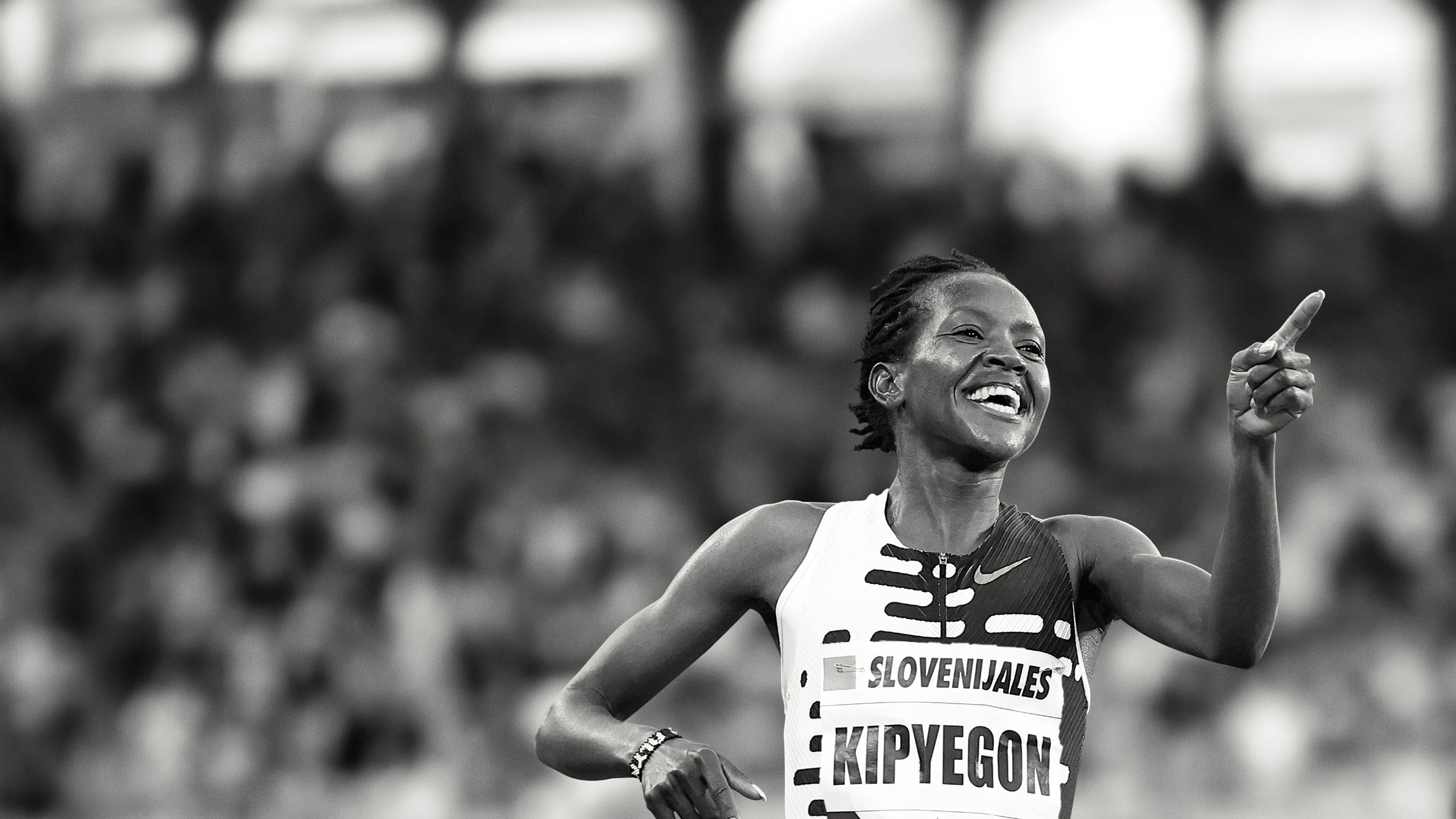 Black and white photo of Faith Kipyegon with a celebration smile and pointing a finger forward. She wears a patterned Nike running top, and there is a crowd behind her.