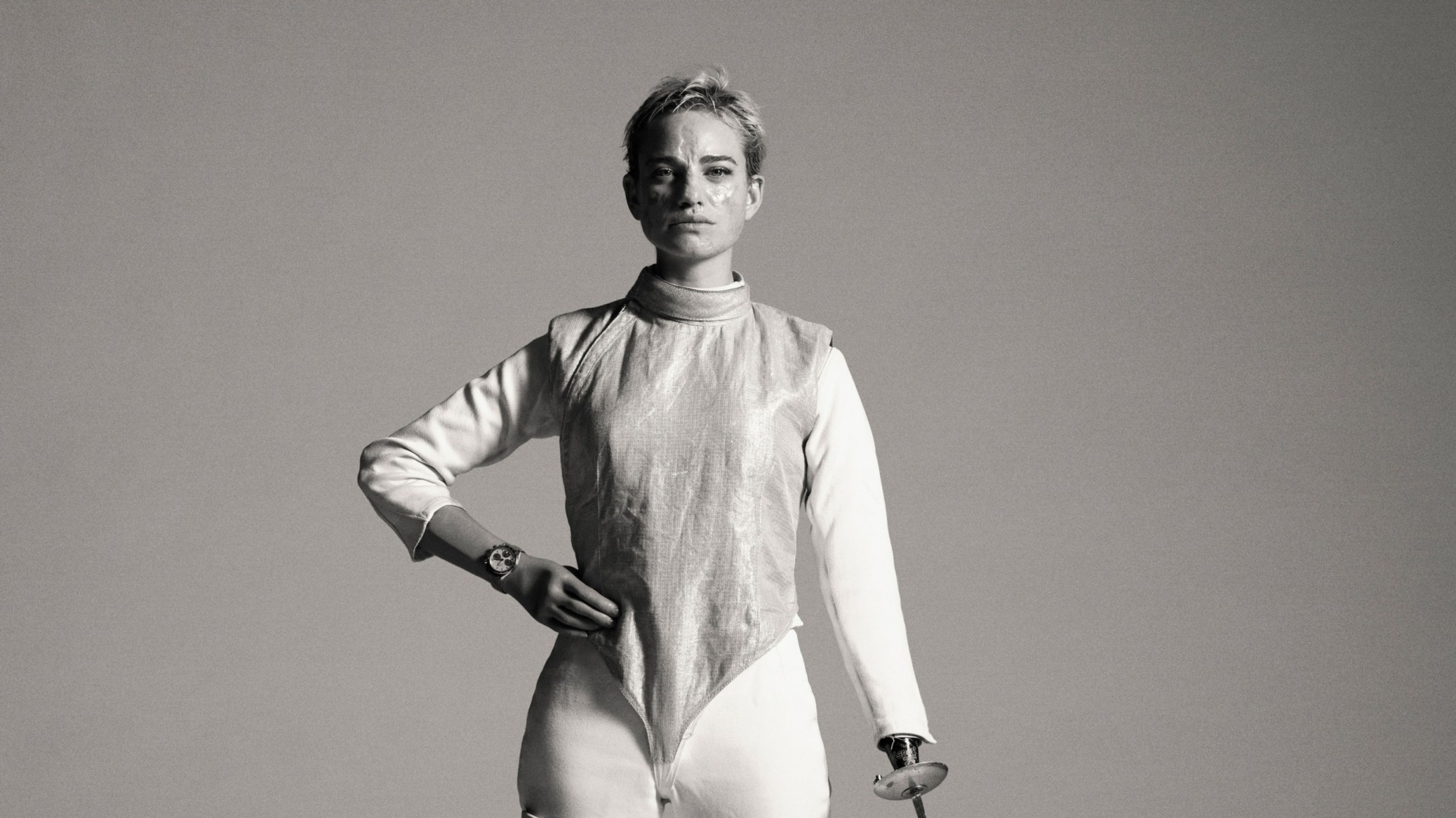 Black and white full body portrait of Bebe Vio, holding one hand to her hip and using her other arm to touch her fencing foil tip to the ground. Bebe looks unsmiling directly at the camera, as she stands in her fencing suit, standing on two prosthetic legs.