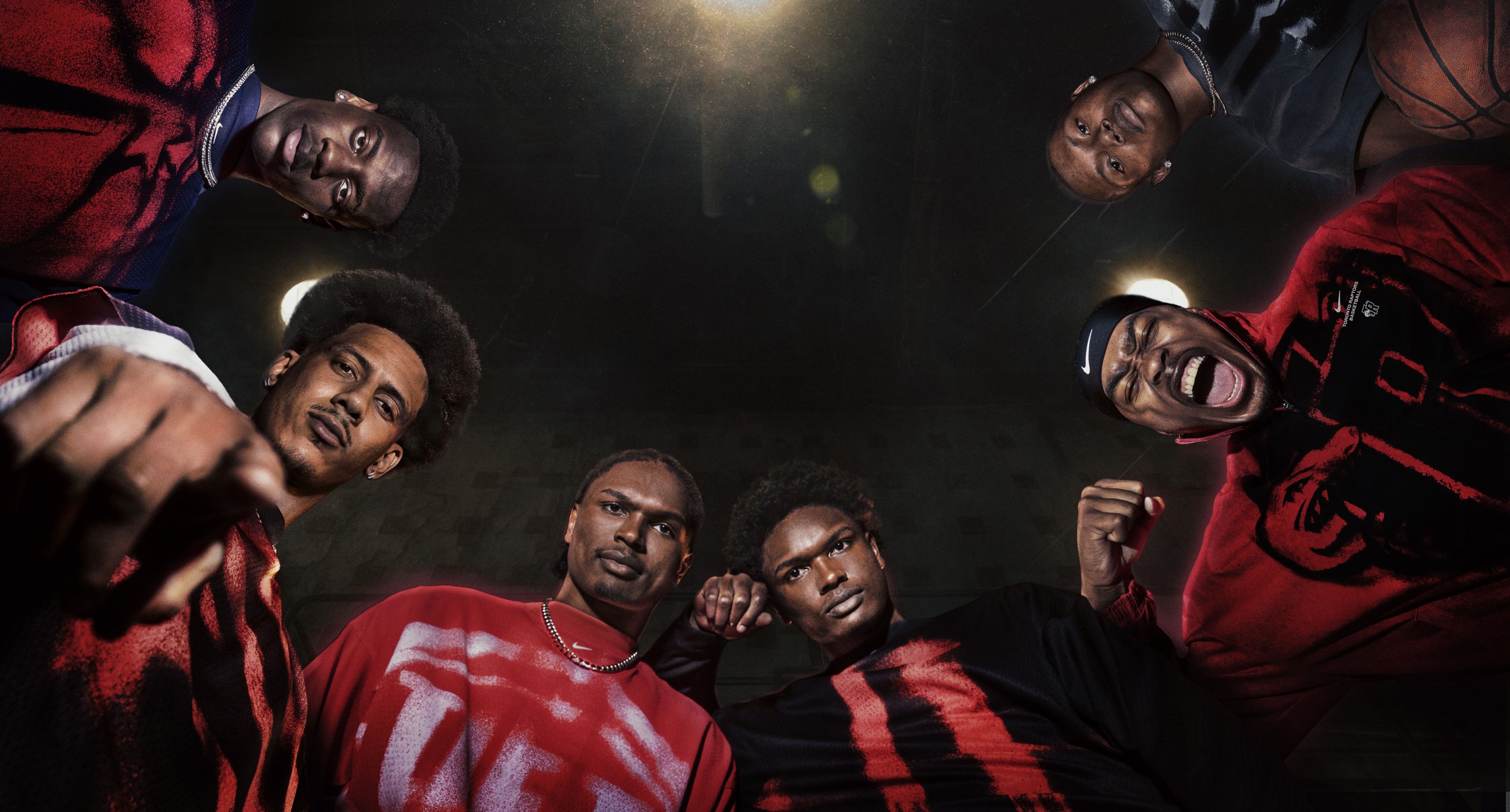 A group of people look down at the camera while wearing pieces from Nike's NBA Standard Issue Collection. The clothing is mostly red and black; the background is black.