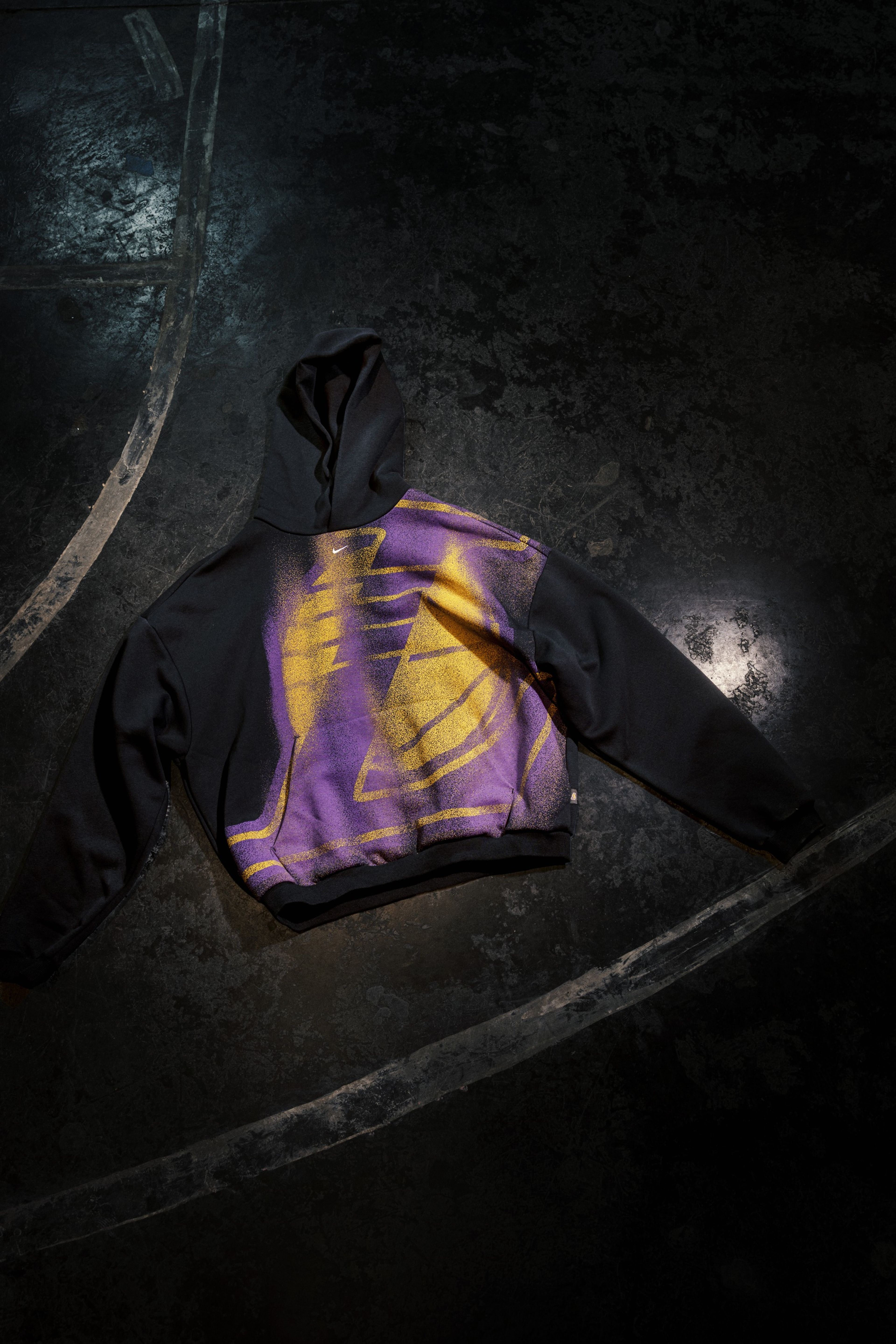 A black hoodie with a large Los Angeles Lakers logo is pictured on a dark basketball court.