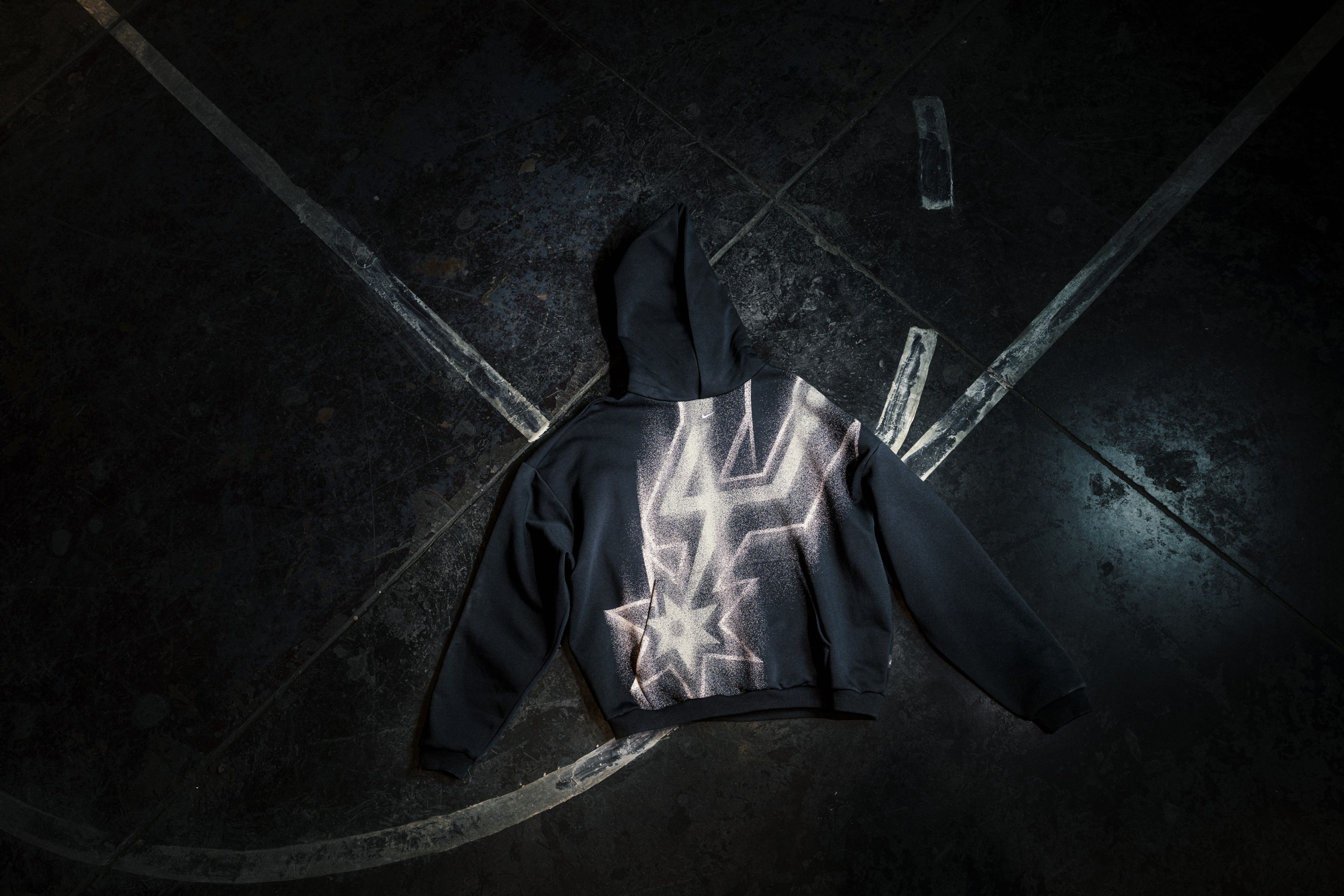 A black hoodie with a large San Antonio Spurs logo is pictured on a dark basketball court.