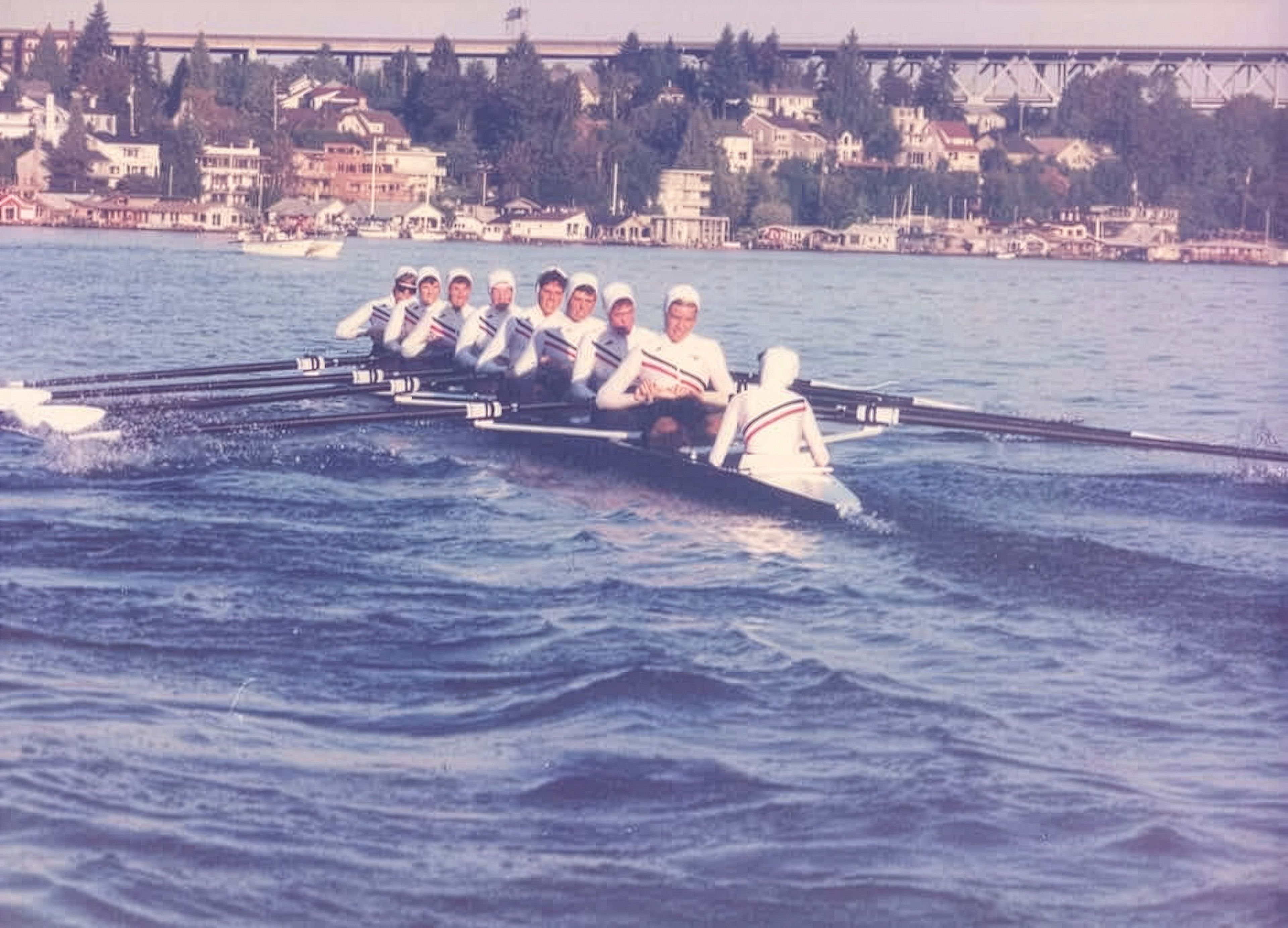 A team of rowers rows away from the camera on a body of water while wearing matching white body suits.