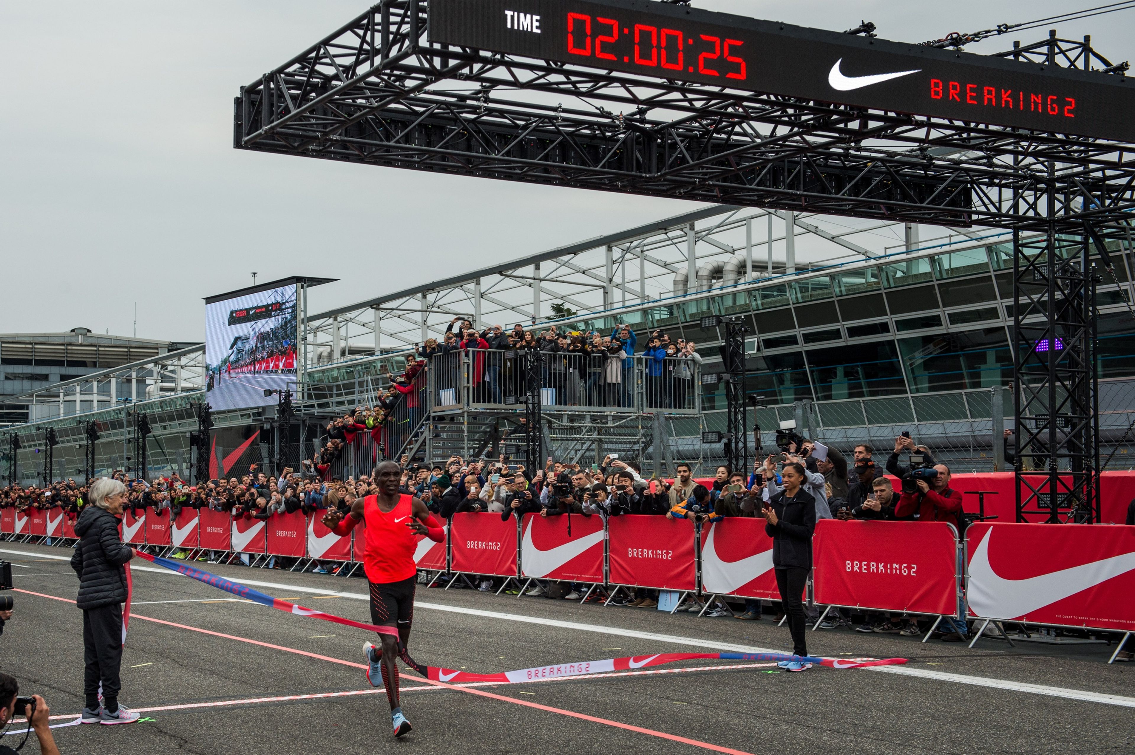 Marathon champion Eliud Kipchoge crosses a finish line. The clock above him reads 02:00:25.