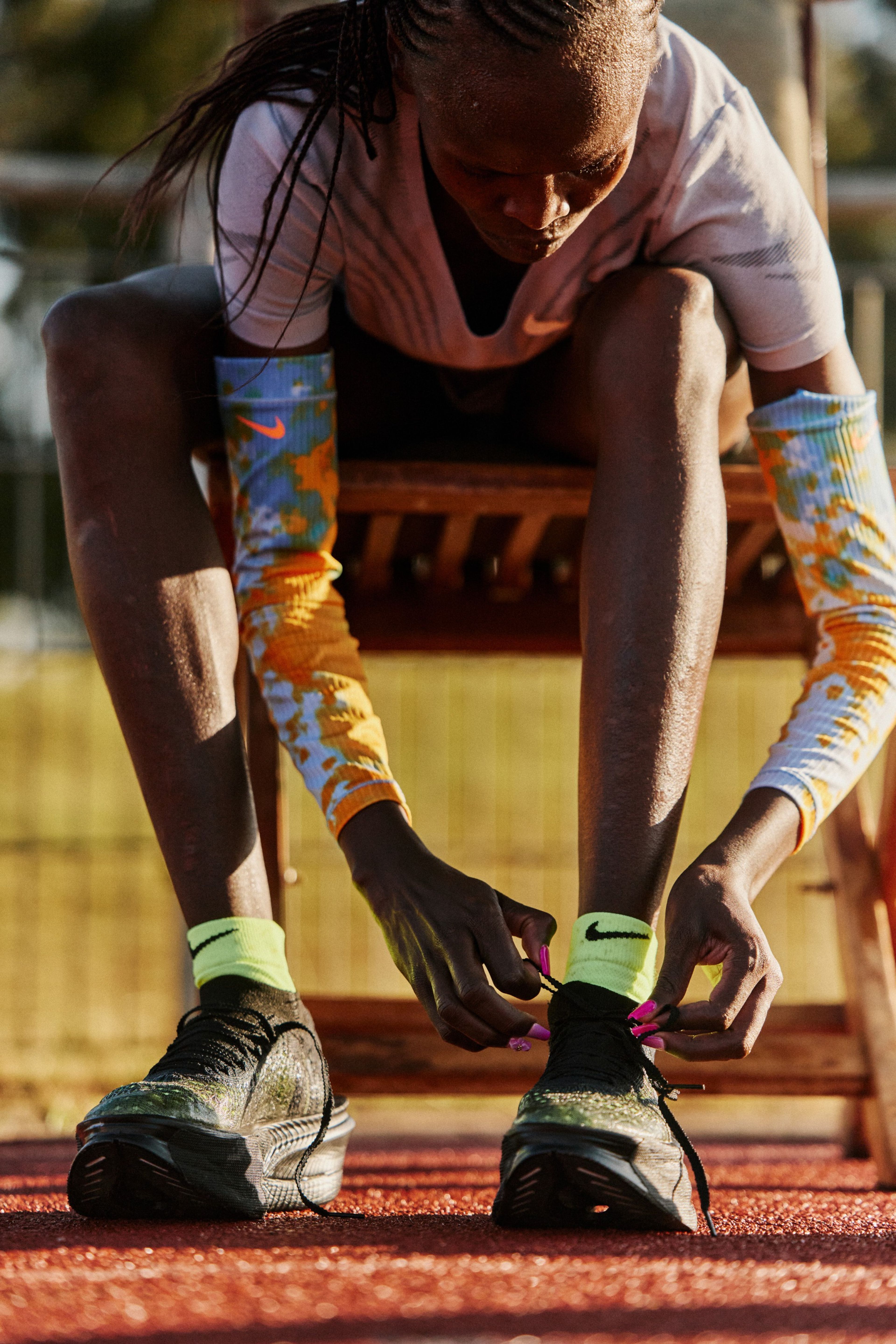 Nike athlete Brigid Kosgei ties her Apex running shoes while seated on a bench at a track.