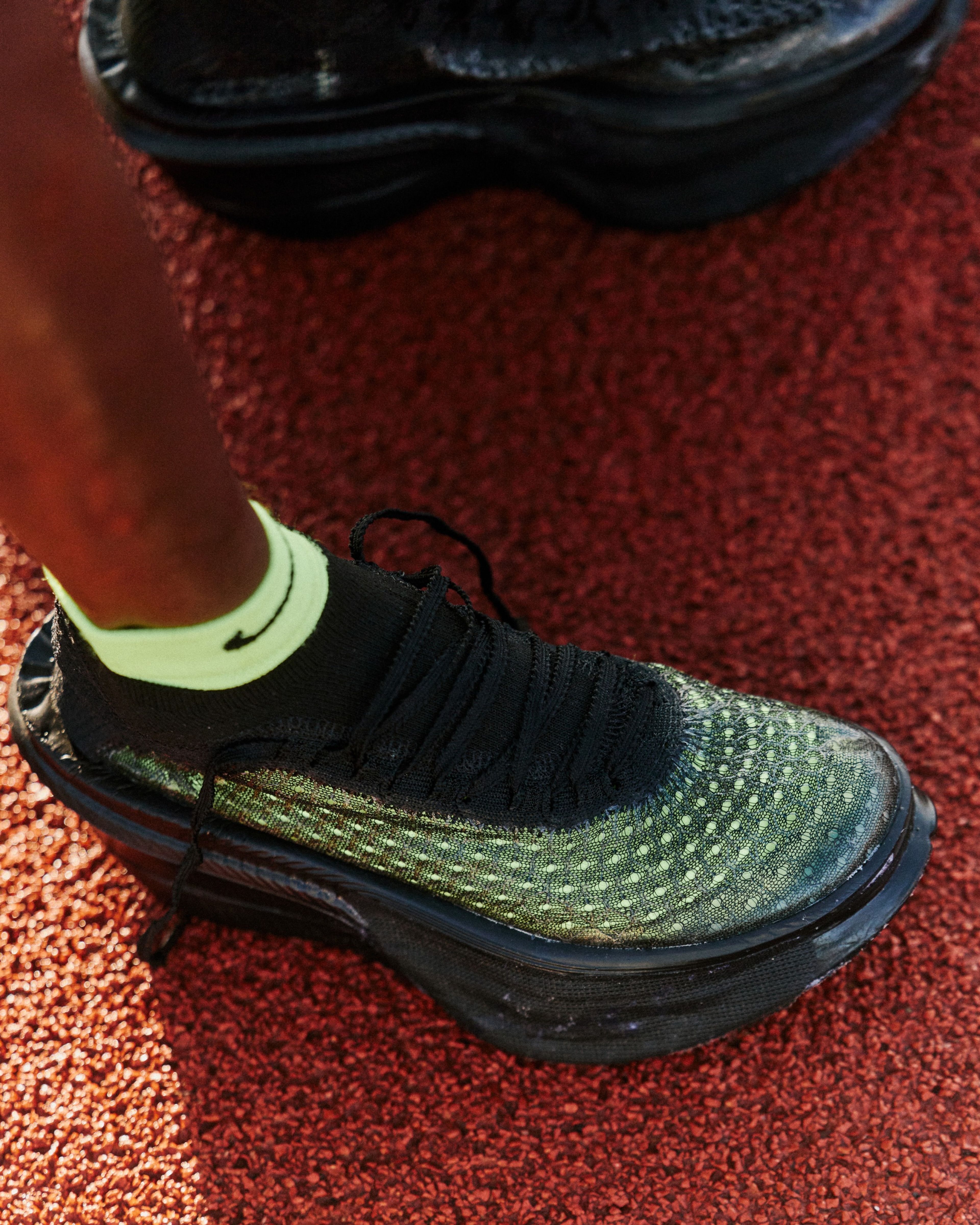 Close-up of a Nike Apex running shoe with a speckled green and black upper, neon green collar, and thick black sole on a red track surface.
