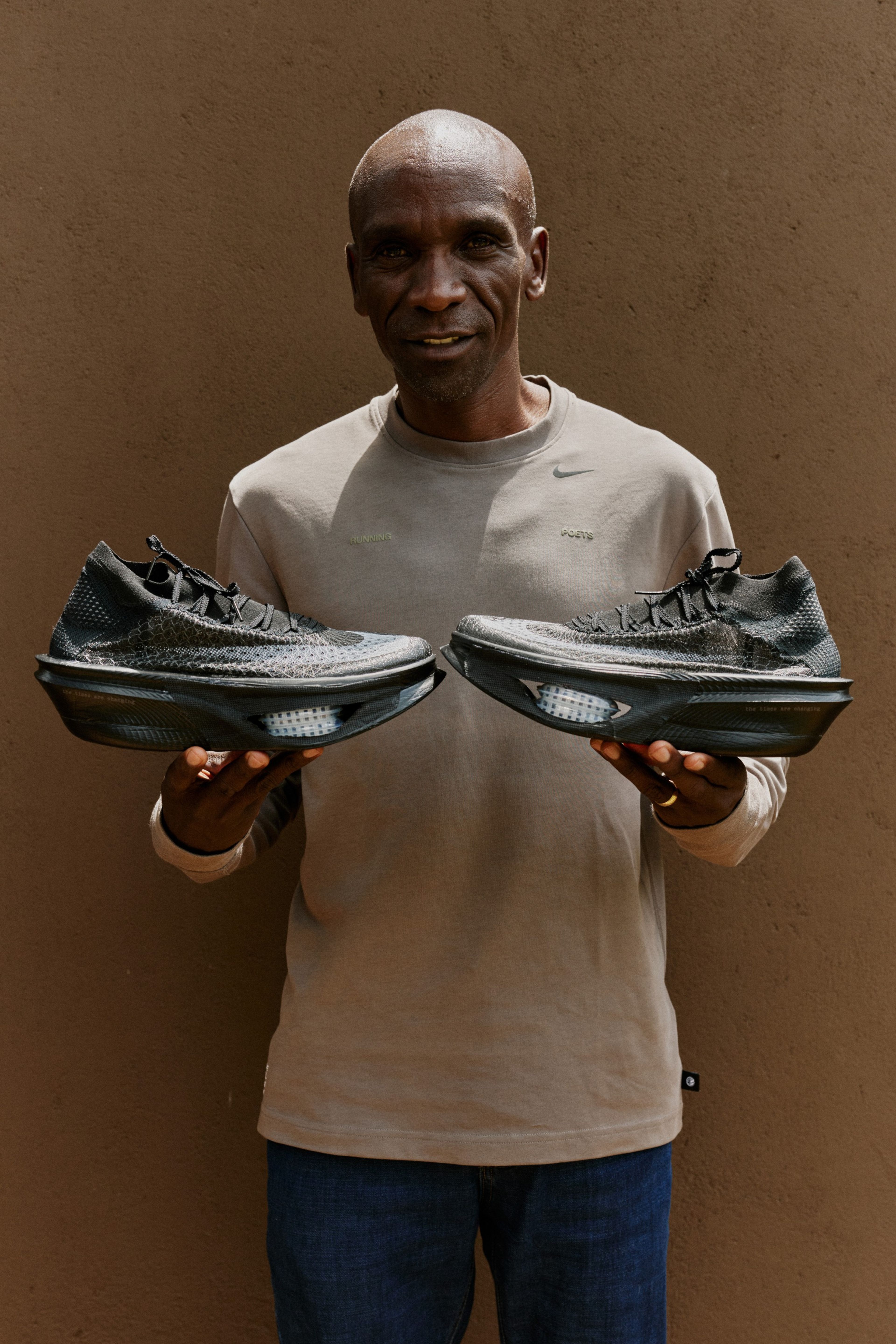 Nike Eliud Kipchoge smiling and holding a pair of Apex running shoes, wearing a beige Nike long sleeve shirt and denim jeans